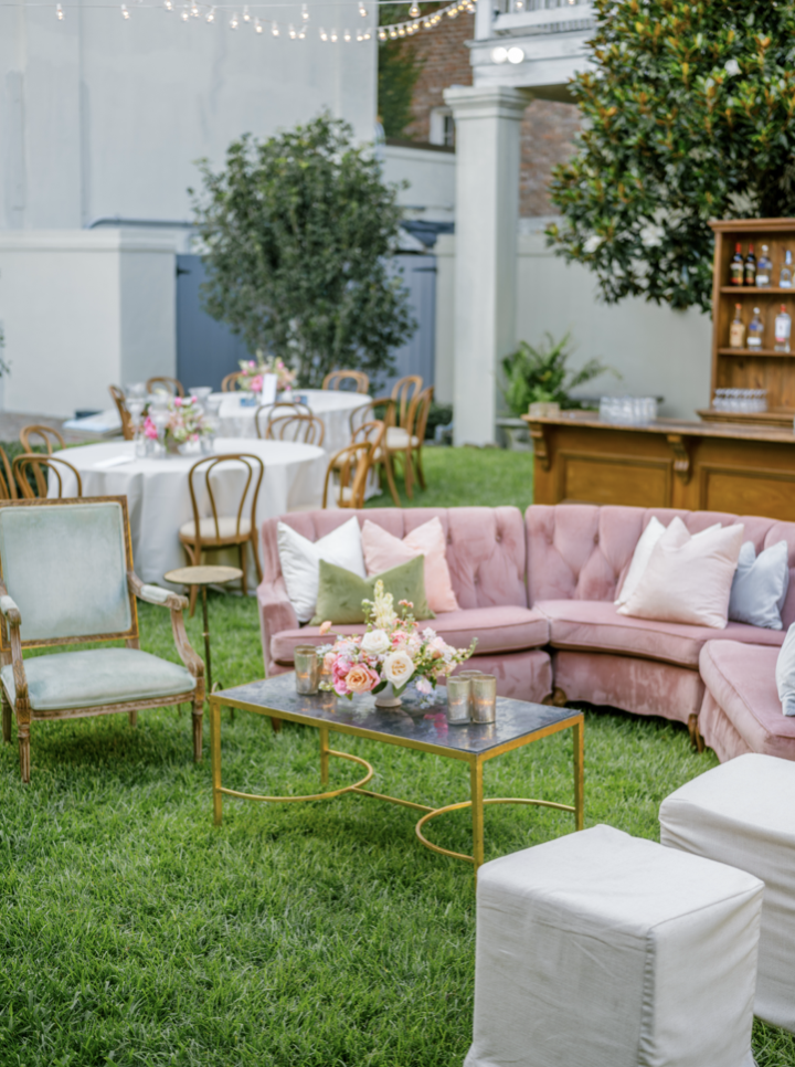 A decorated outdoor garden space with a pink velvet sectional sofa, white and green throw pillows, a pink armchair, a black marble coffee table with a floral arrangement and candles, surrounded by additional chairs and tables with floral centerpieces