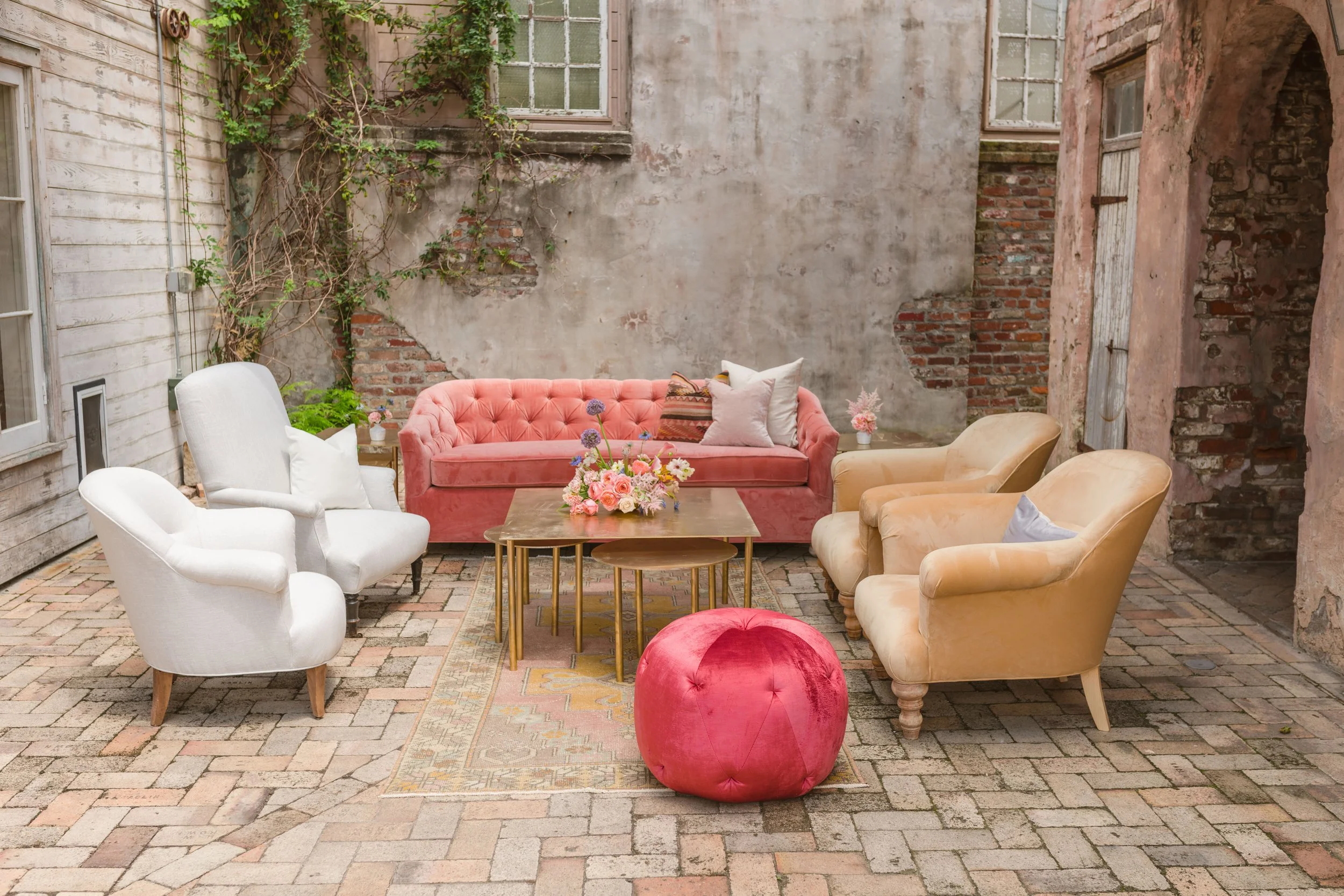 Outdoor patio area with vintage-style furniture including a pink velvet sofa, white and beige armchairs, a pink velvet pouf, and a flower arrangement on a wooden table. The backdrop features a worn brick and stucco wall with greenery growing on it an