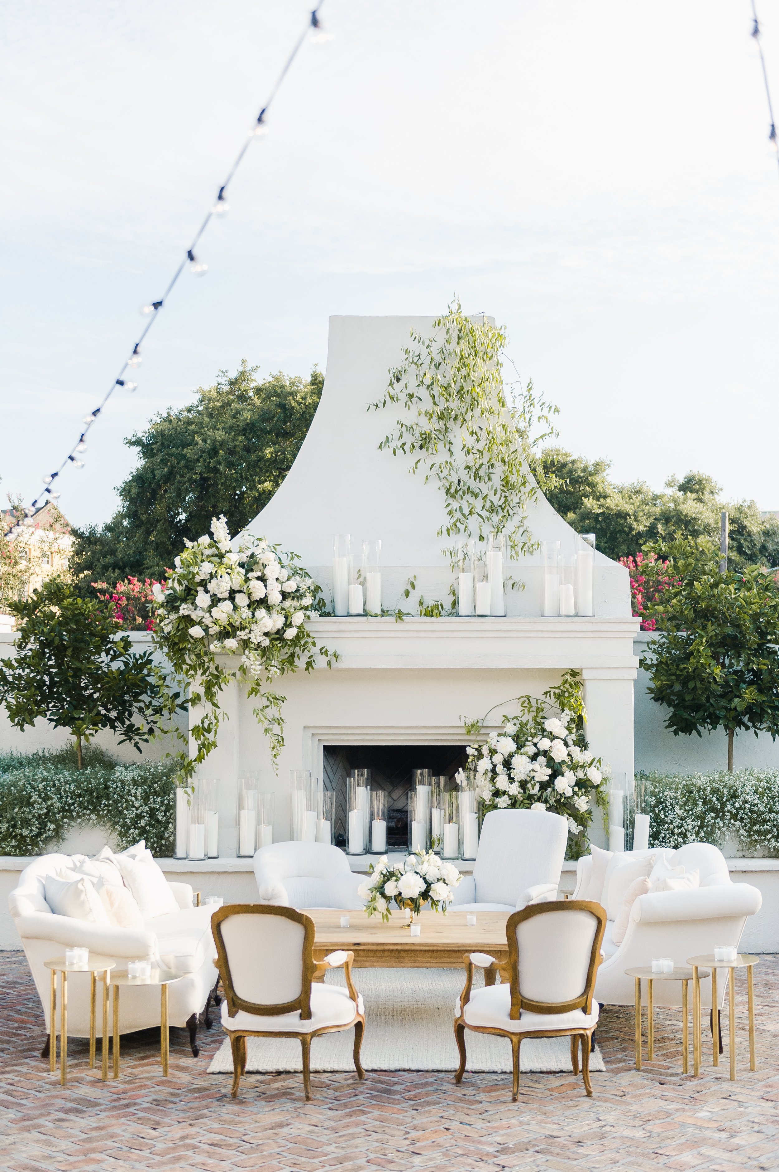 Outdoor wedding or event setup with white chairs, floral arrangements, a white fireplace with candles, and a decorated canopy or backdrop with greenery. Brick patio flooring and trees in the background.