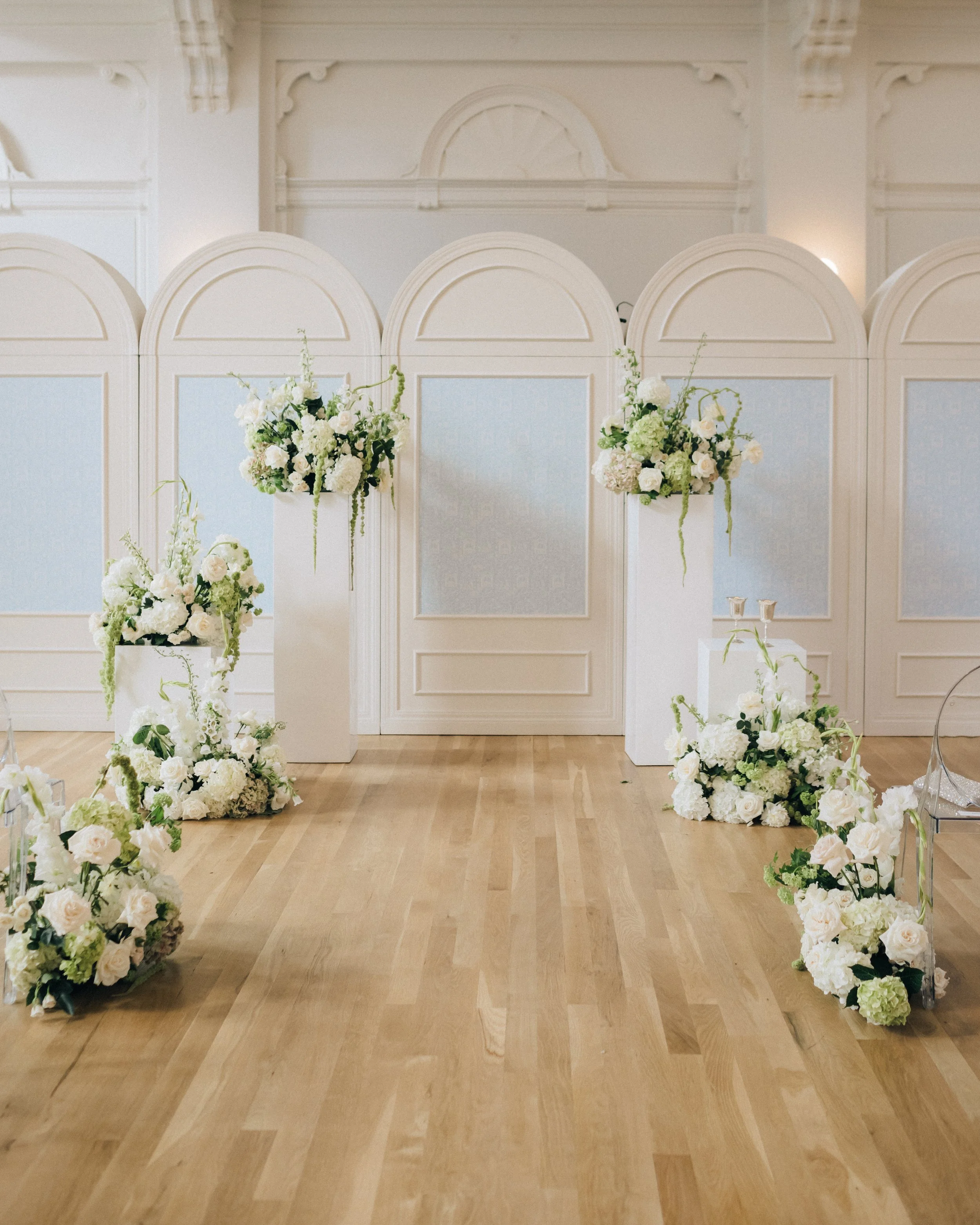 Elegant wedding ceremony setup with white floral arrangements on pedestal stands and flower-strewn aisle on a wooden floor, with an ornate white backdrop.