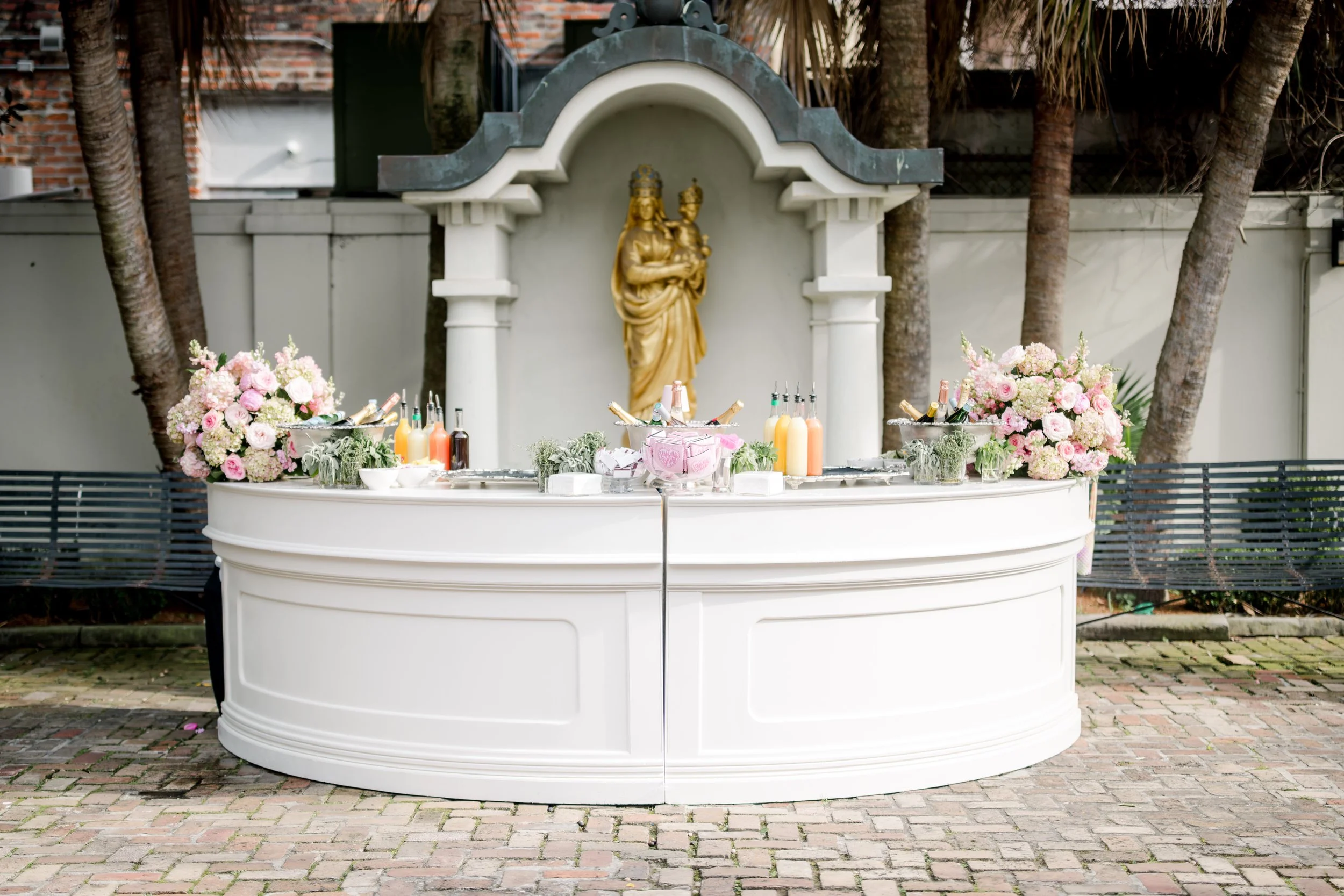 A white bar counter with pink and white floral arrangements on each side, bottles and drinks in the middle, and a gold religious statue with a small child in front of a small semi-circular arch, set outdoors with trees and a brick building in the bac