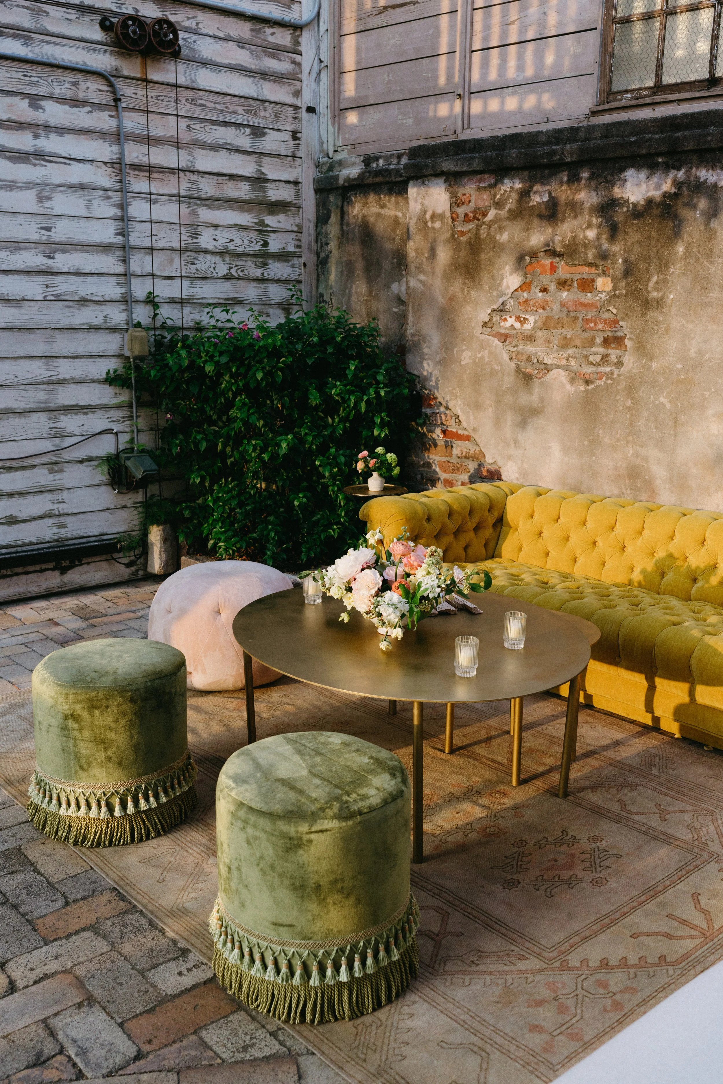 Outdoor seating area with a yellow velvet couch, green velvet stools, floral centerpiece on a round table, and a vintage rug on brick ground. Behind the seating, there is a weathered brick and concrete wall with peeling paint and greenery in the corn