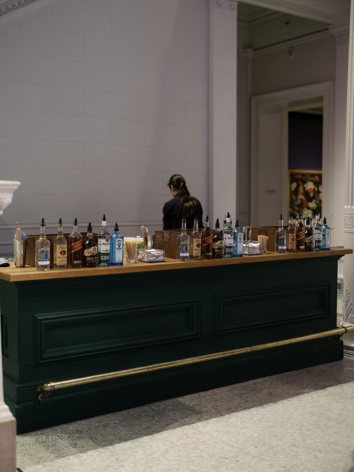 Bar with a row of liquor bottles and barware, behind which a woman in black is standing with her back to the camera in a formal setting.