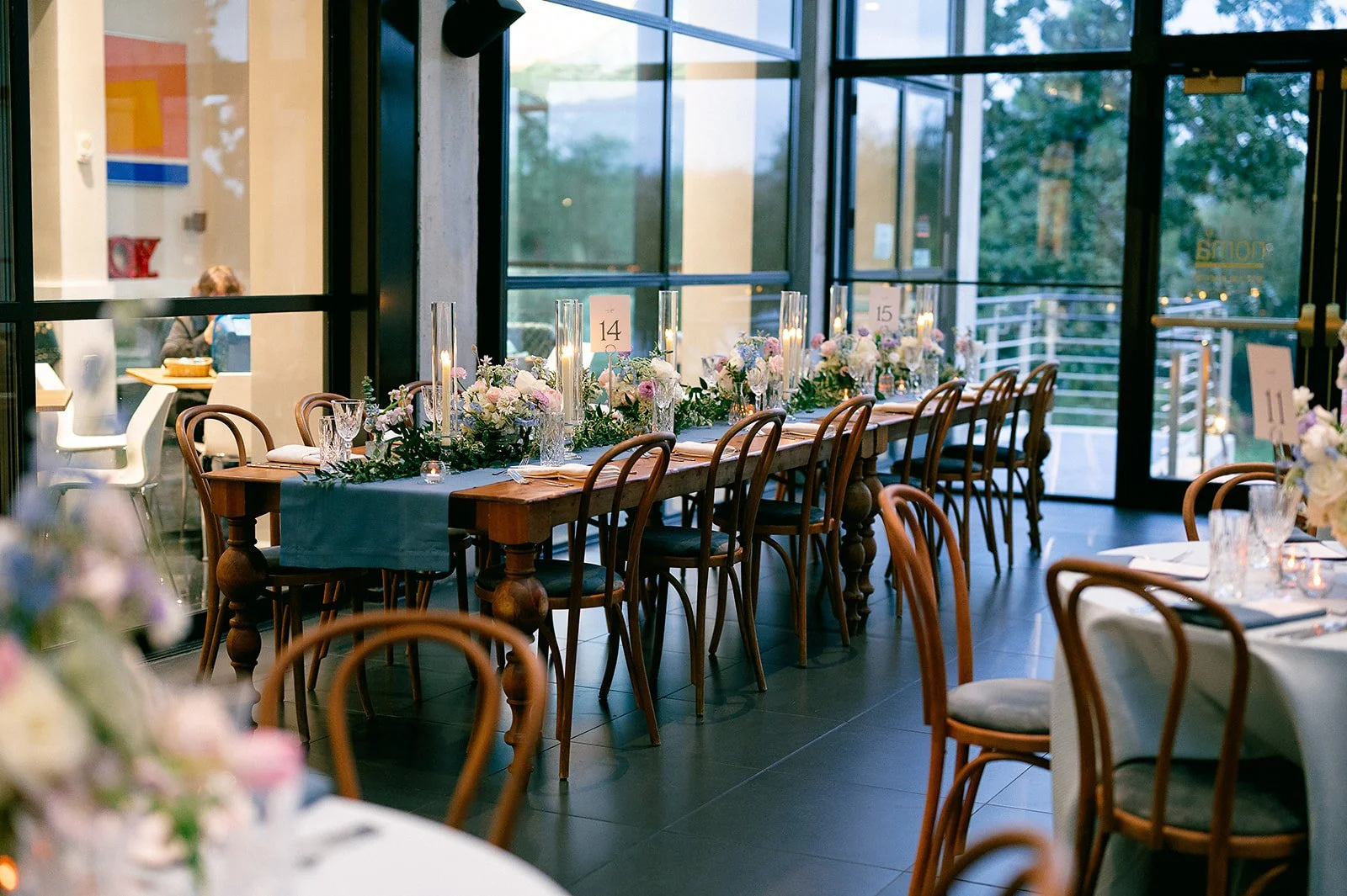Elegant banquet table set for a wedding reception with floral centerpieces, tall candles, and table numbers, inside a venue with large glass windows and outdoor greenery.