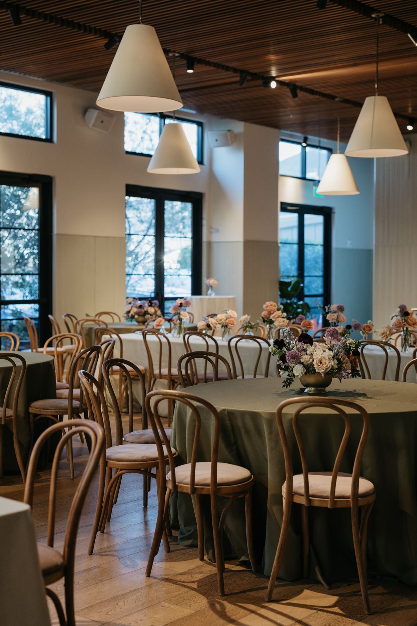 Indoor event space with round tables covered in green tablecloths, decorated with floral centerpieces, and surrounded by wooden chairs. Large windows let in natural light, and white cone pendant lights hang from a wooden-paneled ceiling.