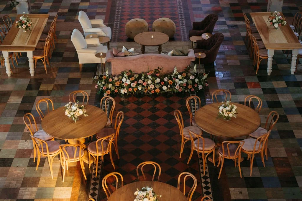 An elegant indoor event setup with round wooden tables and chairs, floral centerpieces, a decorated seating area with armchairs, and floral arrangements, all on a patterned tiled floor.