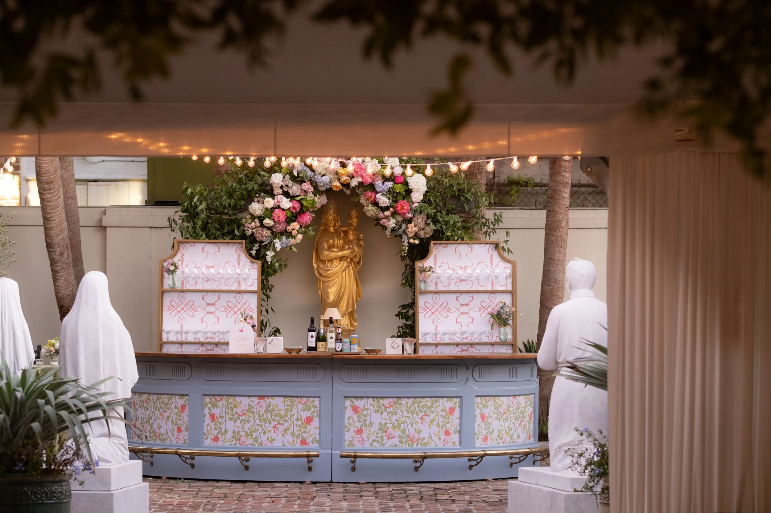Outdoor wedding bar setup with floral decorations and gold statue, surrounded by trees and string lights.