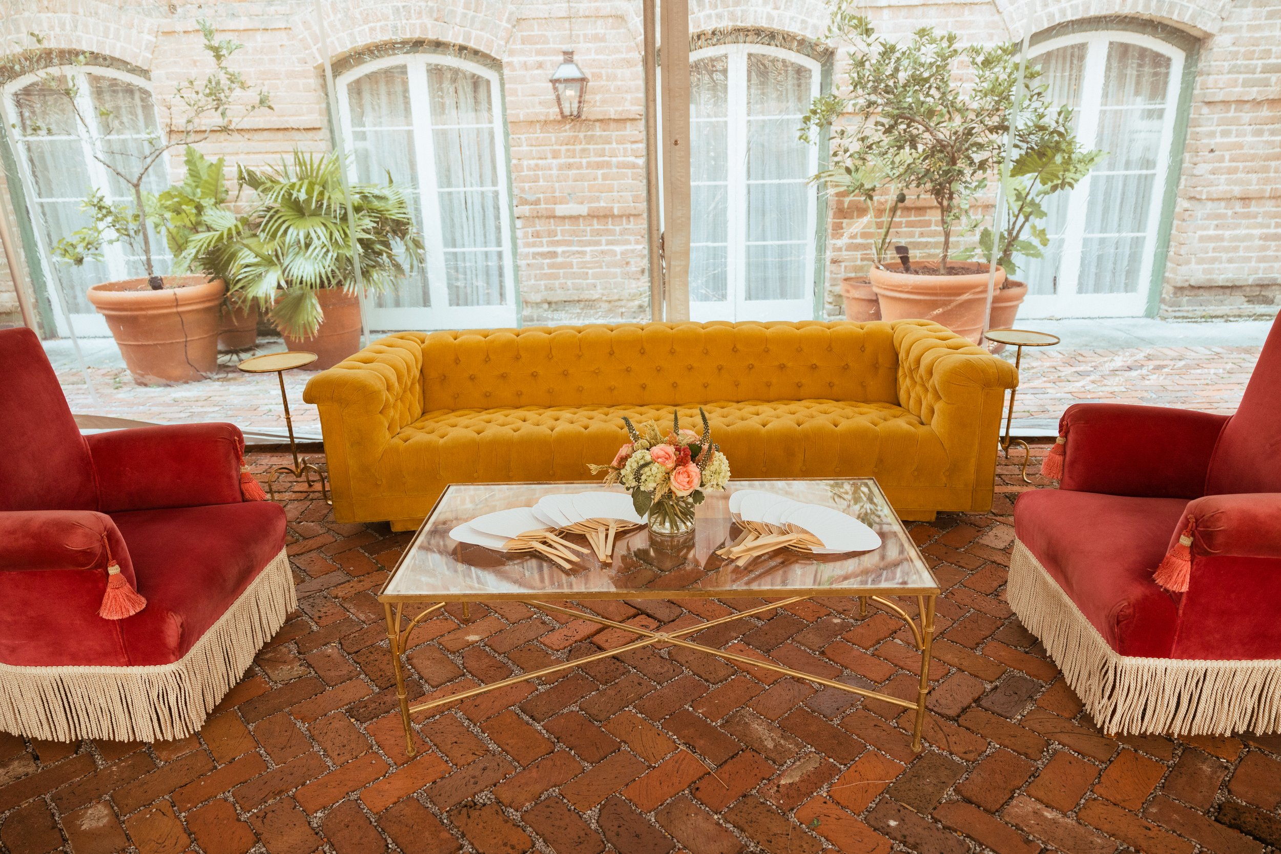 Interior of a room with a glass wall showing outdoor potted plants and brick flooring, furnished with a yellow tufted sofa, two red armchairs with fringed skirts, a floral centerpiece on a marble coffee table, and small side tables.