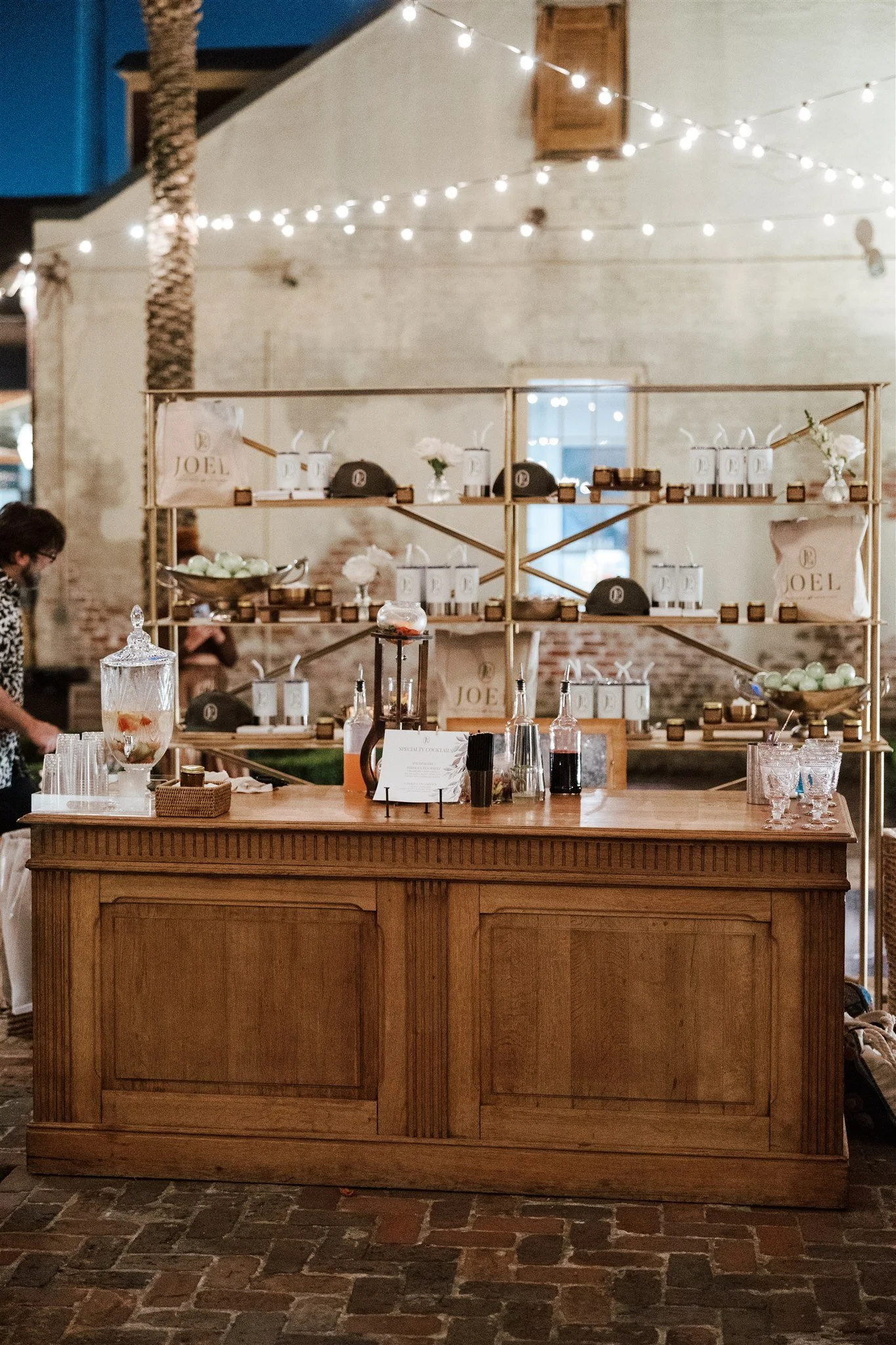 A bar counter made of wood with drinks and glassware on it, set against a brick floor and a wall decorated with warm string lights, and a shelf with various items, including bags, hats, and small decorative objects.
