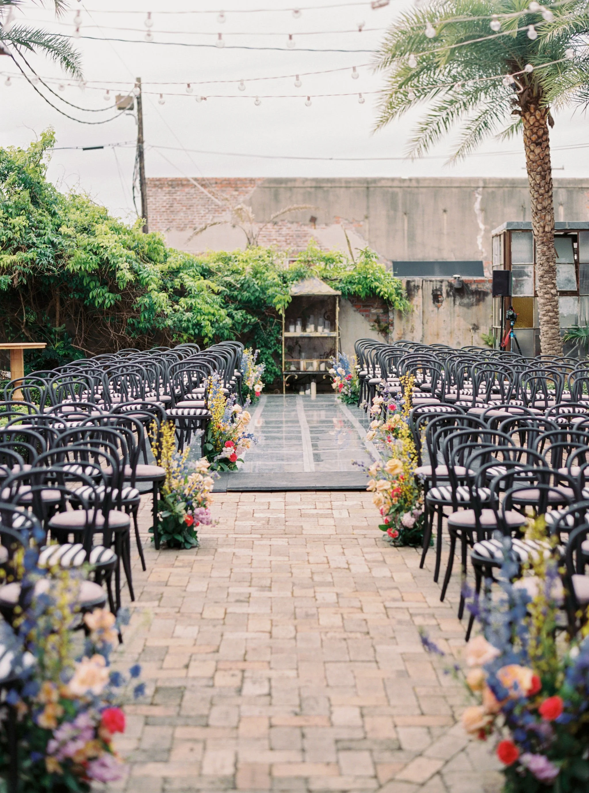 Outdoor wedding ceremony setup with rows of black chairs on a brick walkway, decorated with colorful flowers on either side, leading to an altar. String lights hang overhead, surrounded by greenery and palm trees, with an urban brick wall in the back