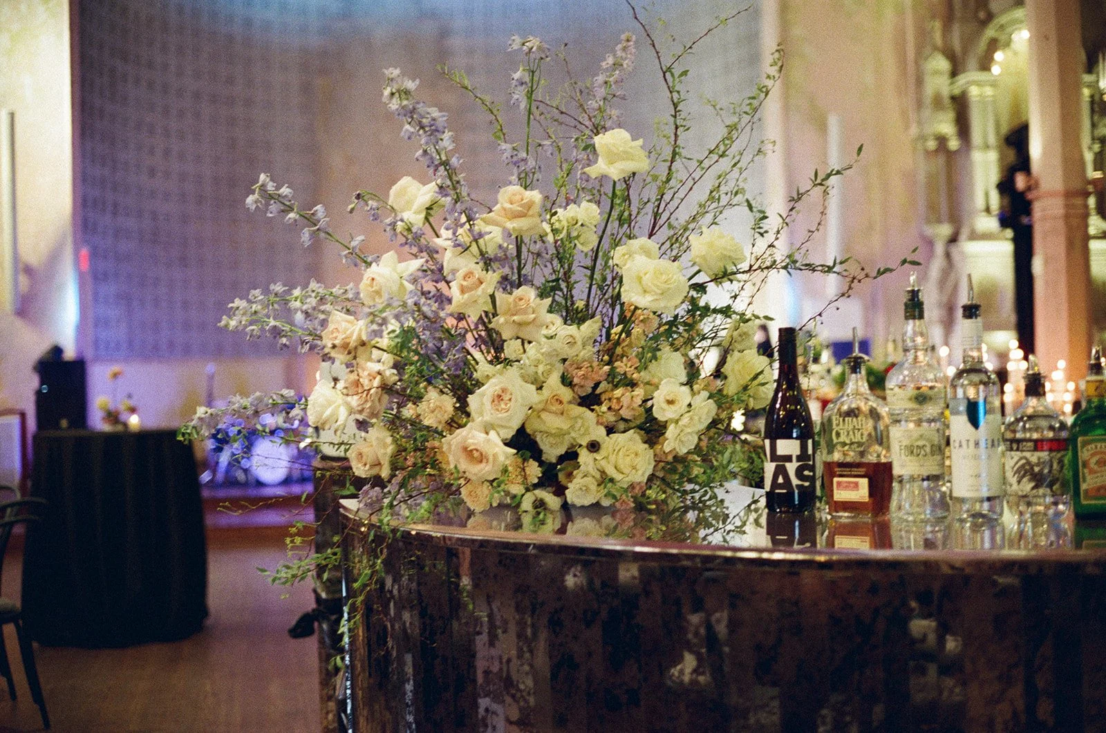 A floral centerpiece with white and pastel-colored flowers on a bar counter with liquor bottles in an elegant, warmly lit interior.