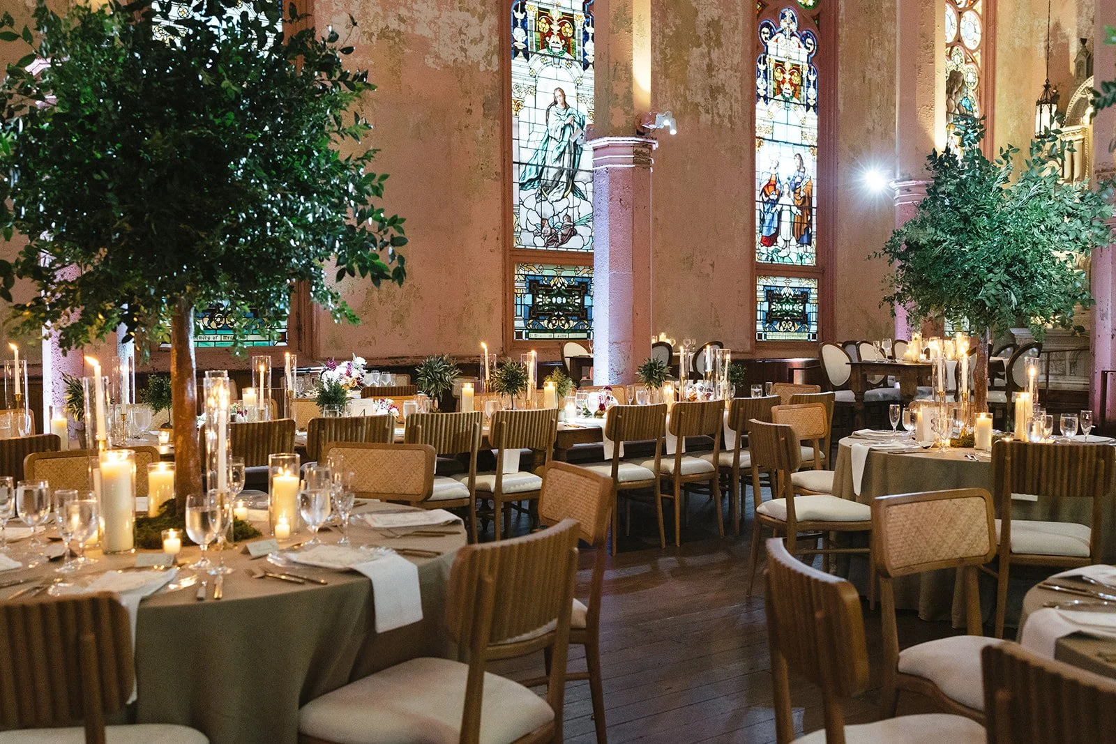 Elegant dining setup inside a church with stained glass windows, surrounded by tables with candles, glassware, and floral centerpieces, and decorated with potted trees.