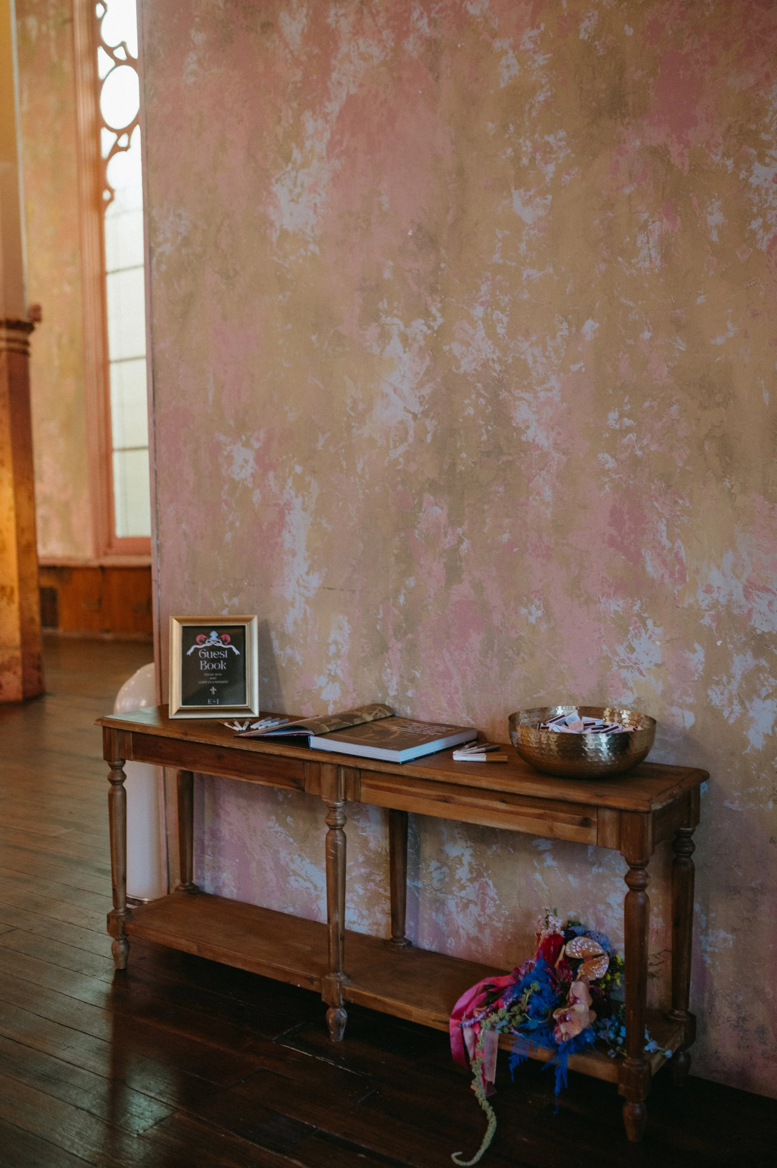 A wooden console table against a pink textured wall. On top of the table are some books, a framed guest book, and a decorative bowl. On the lower shelf of the table, there are some floral leis and ribbons. To the left in the background is a window wi