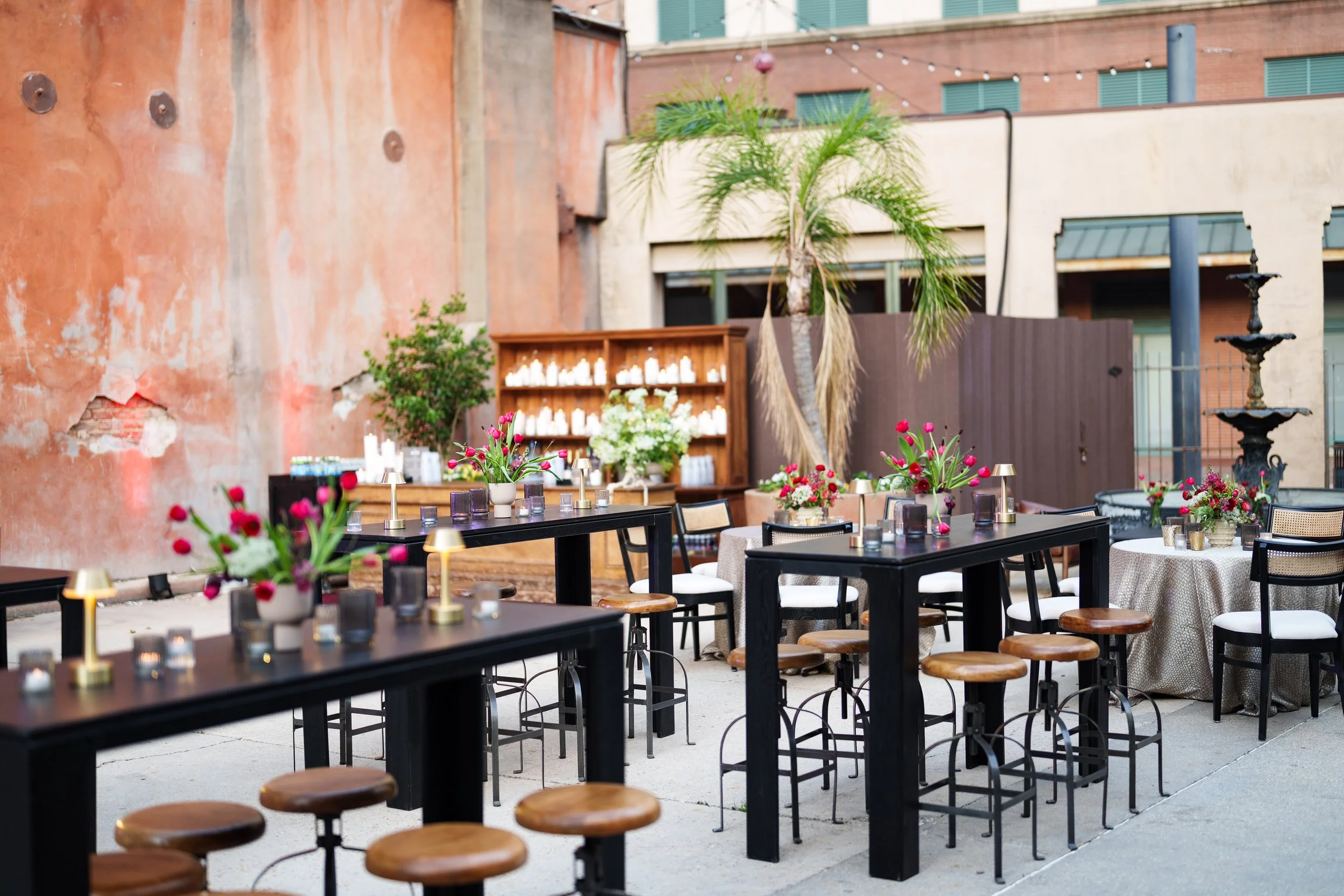 An outdoor patio with tables decorated with flowers and candles, surrounded by chairs and stools, set against a weathered red wall and a modern building.