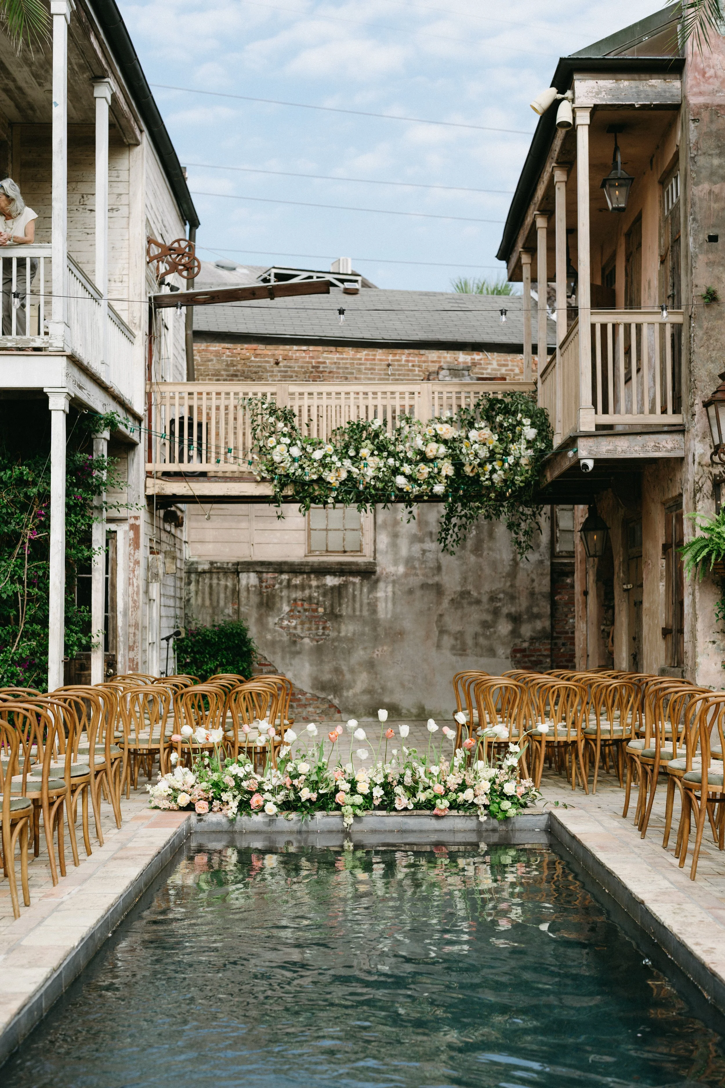 Outdoor courtyard with chairs arranged around a floral centerpiece, a water feature in the foreground, and a backdrop of rustic buildings decorated with flowers and string lights.