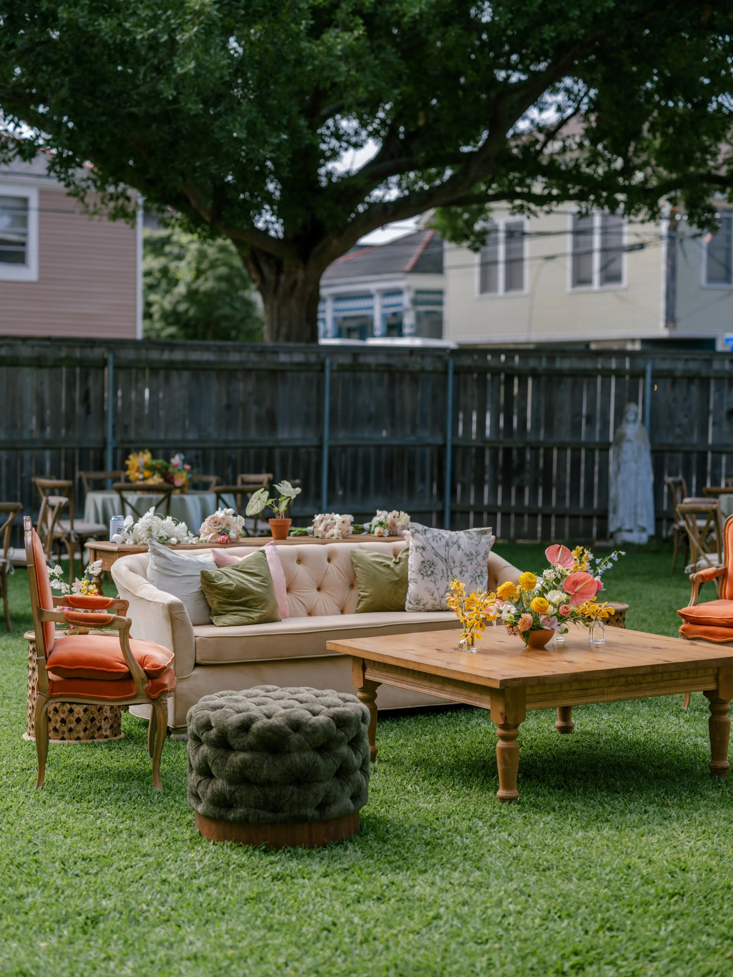 Outdoor backyard decorated with vintage furniture, including a beige tufted sofa with patterned and velvet pillows, surrounded by floral arrangements, a wooden coffee table, and a velvet pouf, with a large tree and neighboring houses in the backgroun