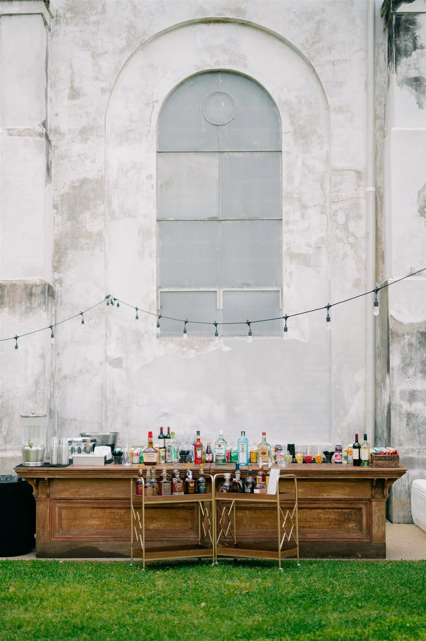 A bar setup outdoors with a long wooden counter topped with various bottles of alcohol, mixers, and bartending tools. There are metal barstools in front and string lights hanging above. The background features a large, weathered concrete wall with a 