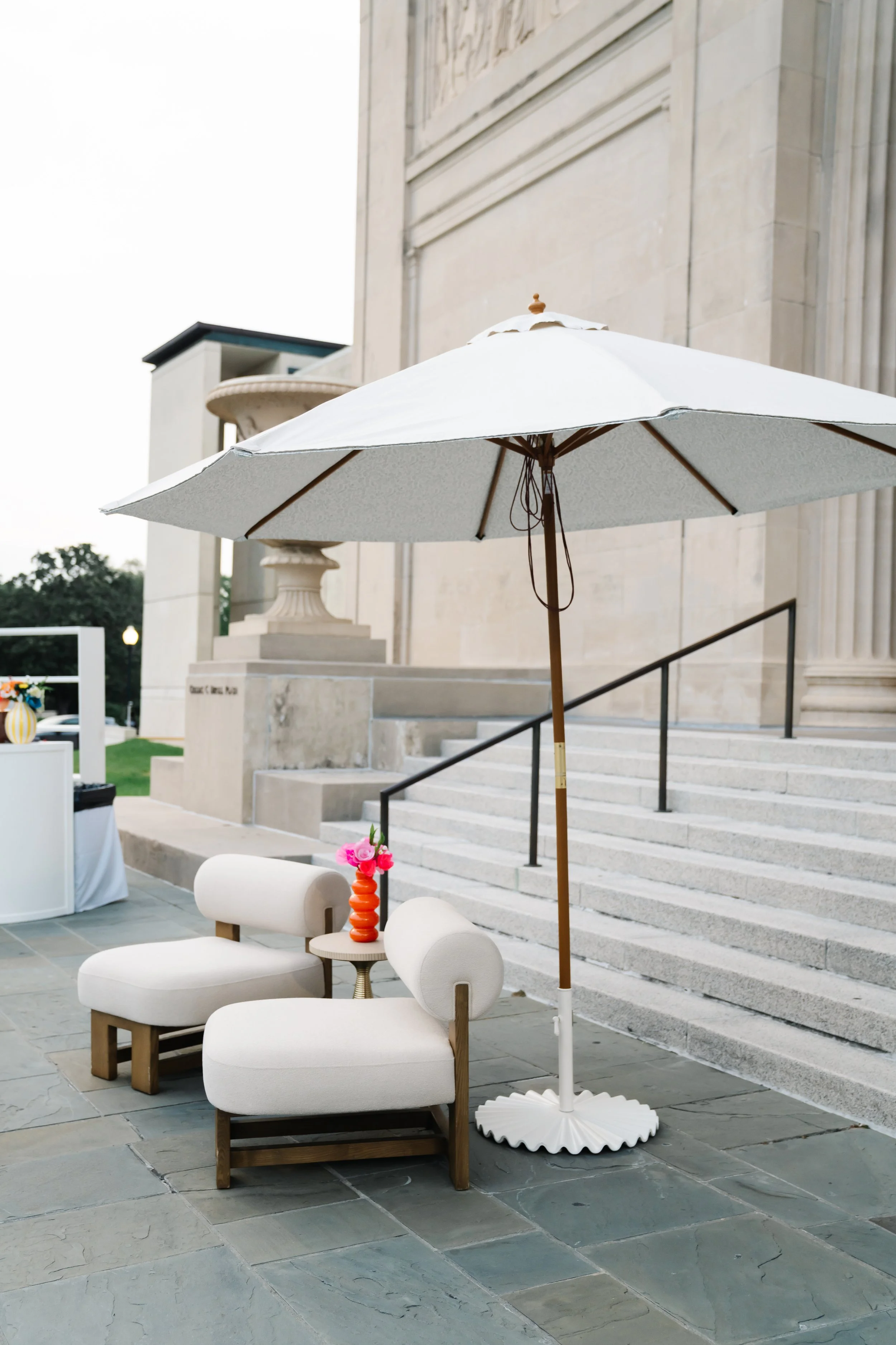 Outdoor seating area with two modern white lounge chairs, a small side table with a decorative orange vase containing pink flowers, and a large white patio umbrella. In the background are stone steps leading up to a building with beige stone walls an