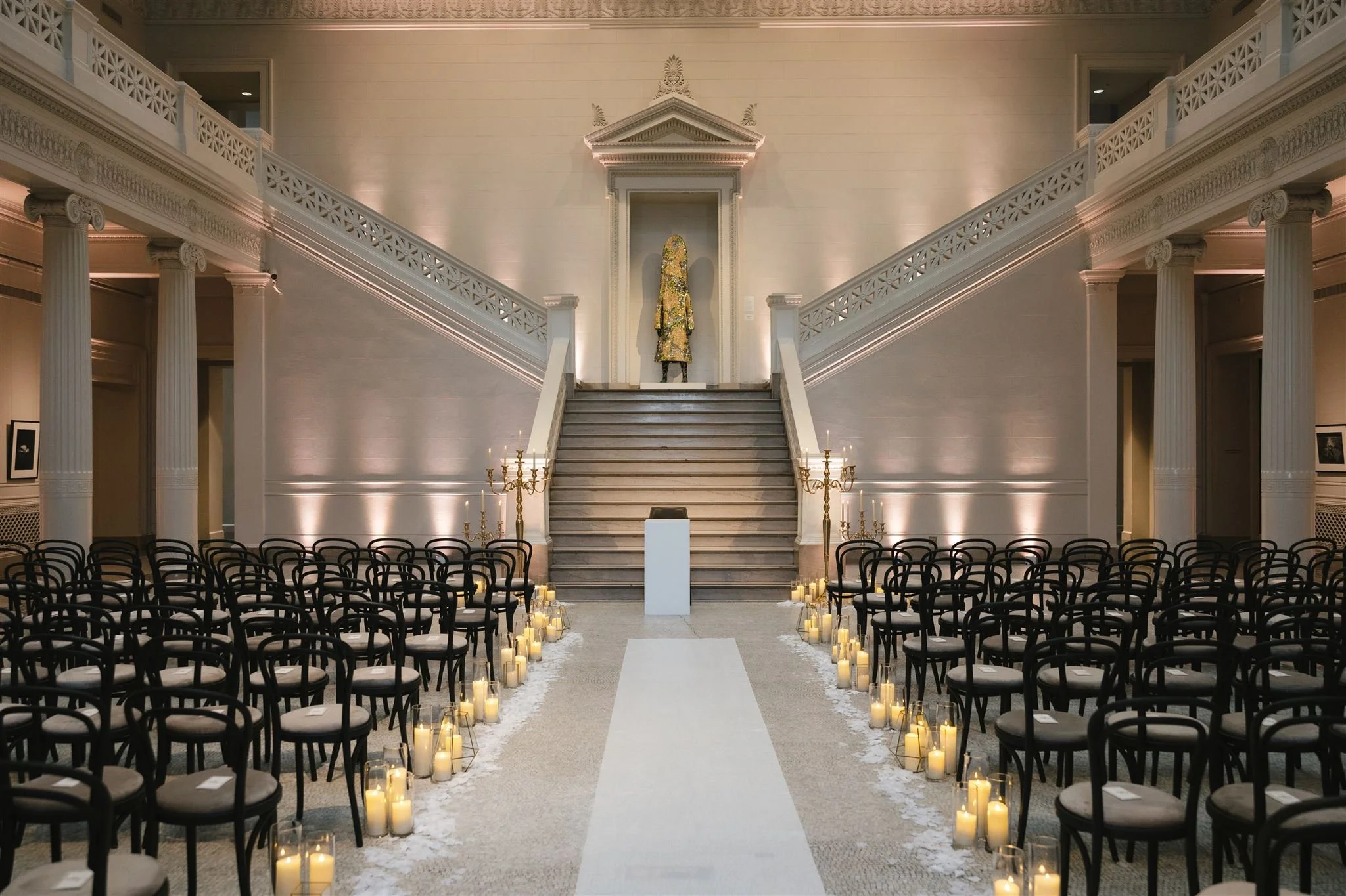 Elegant indoor wedding ceremony setup with chairs arranged in rows on either side of a white aisle. Candles in glass containers line the aisle, leading to a staircase with a statue dressed in decorative clothing at the top, illuminated by soft lighti