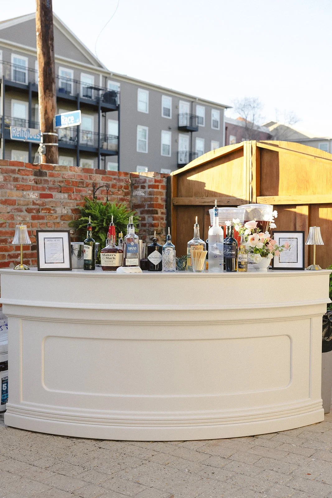 Outdoor bar setup with various bottles of alcohol, framed menus, and a flower arrangement, positioned in front of a brick wall and wooden fence.