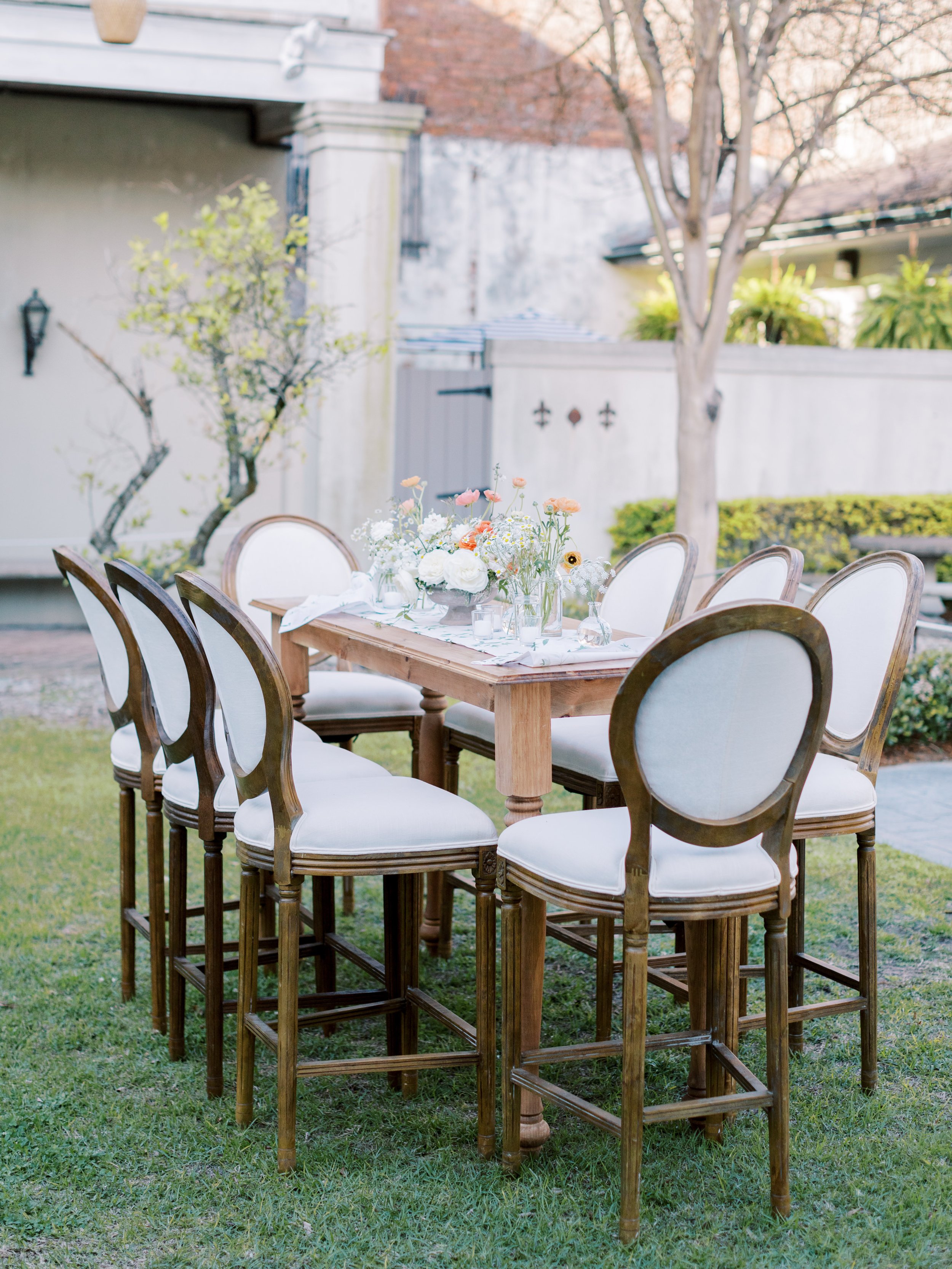 A wooden outdoor dining table with a floral centerpiece and eight white upholstered chairs with wooden frames set on green grass in a backyard garden.