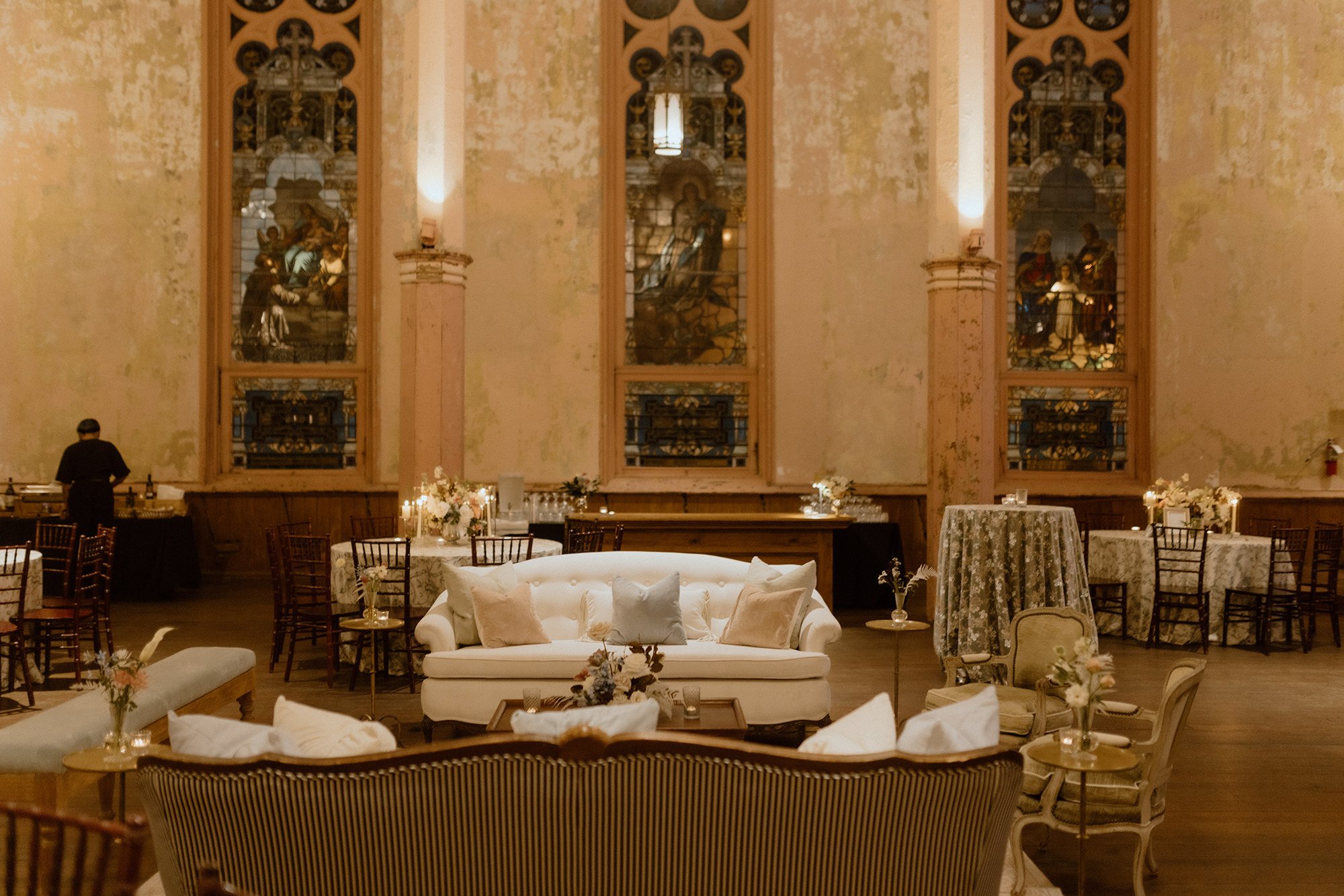 Elegant event venue with soft lighting, decorated tables with floral arrangements, a white tufted sofa with cushions, large stained glass windows depicting religious scenes, and a waiter preparing the tables.