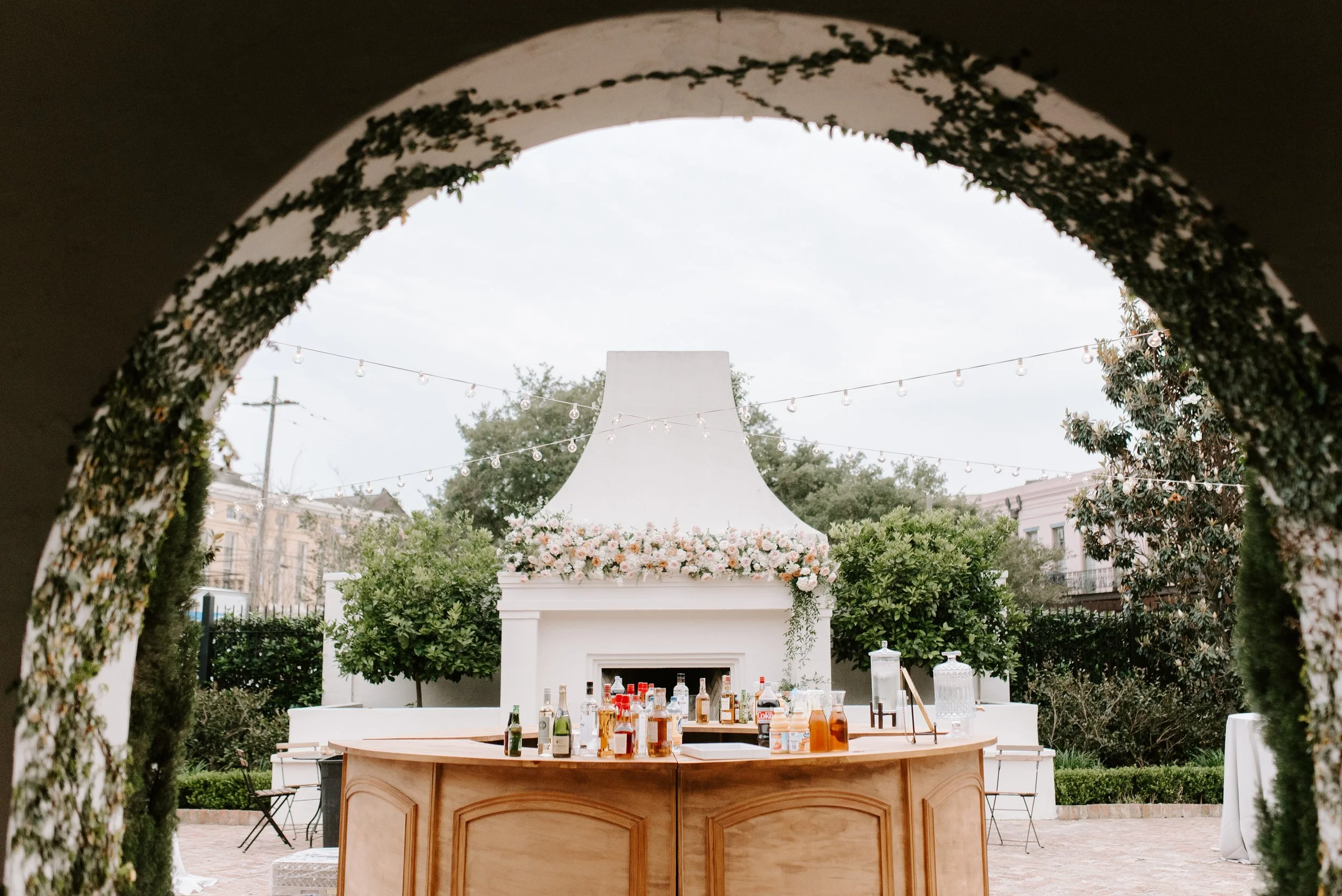 Wedding reception setup outdoors with a bar table in front, decorated with bottles and drinks, under a white floral arch and string lights, with trees and buildings in the background.