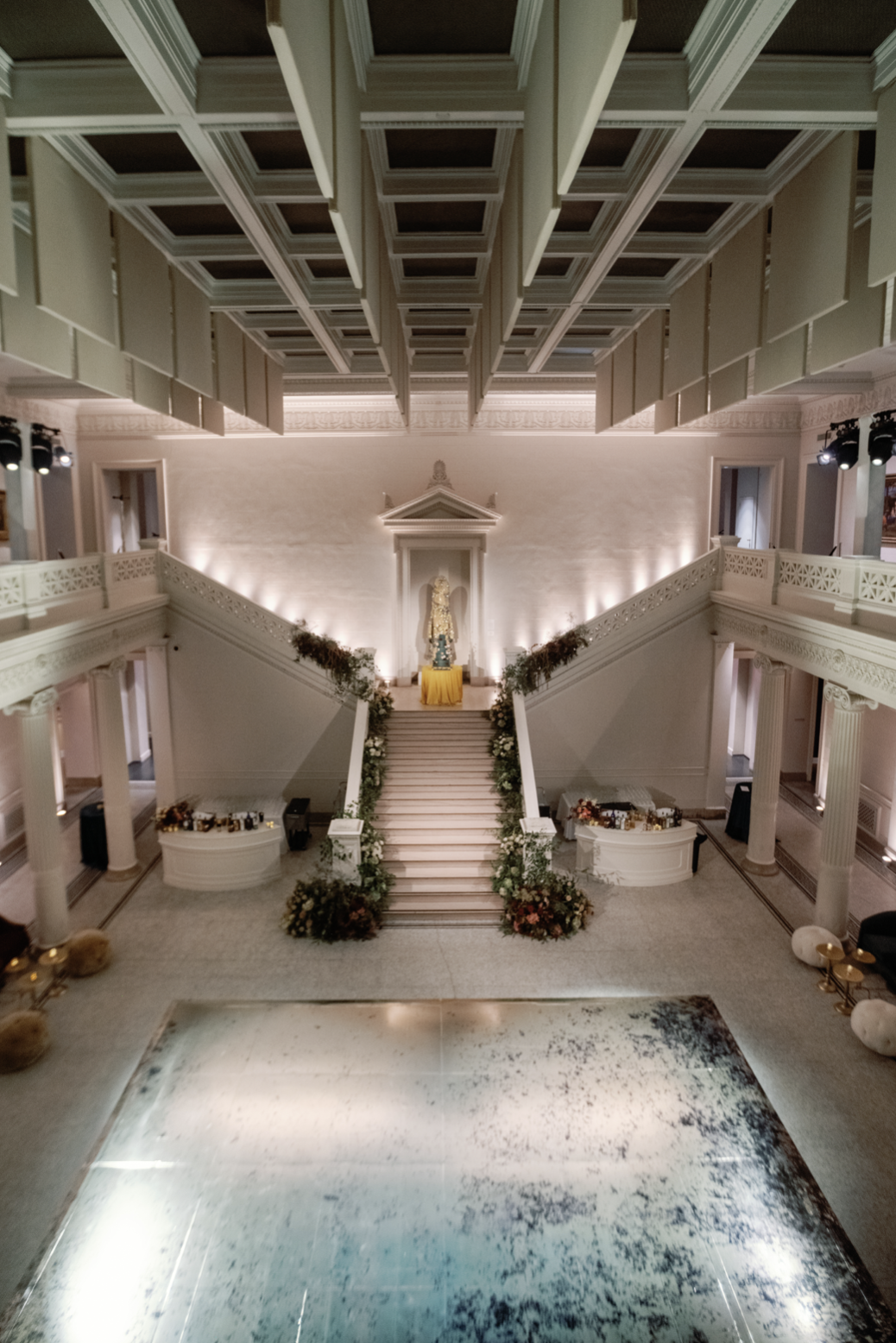 Elegant indoor event space with a grand staircase decorated with flowers leading to a statue on a pedestal. A dance floor is in the foreground, with soft lighting and architectural details.