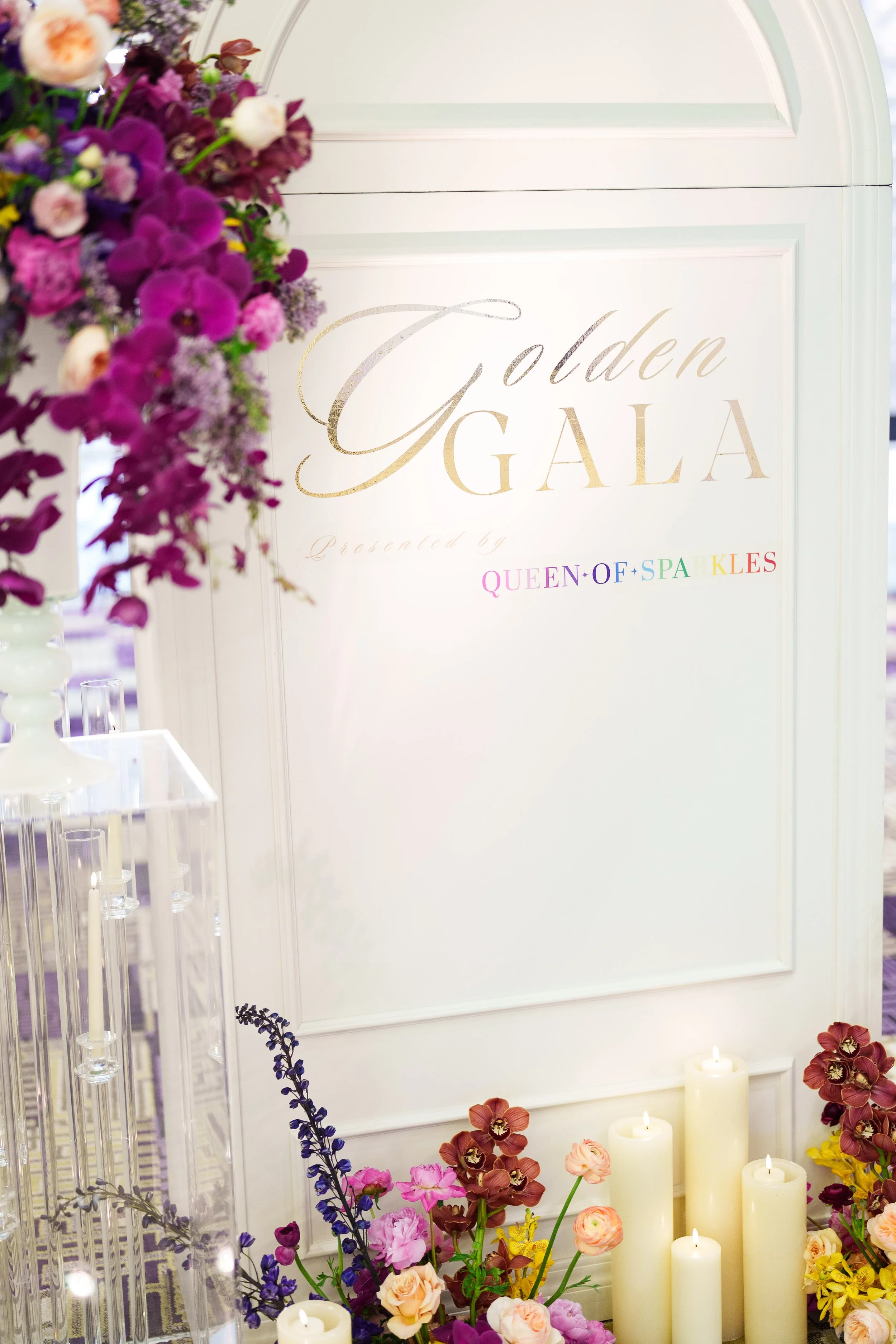 A welcome sign for the Golden Gala event, presented by Queen of Sparkles, with colorful flowers and candles around it.