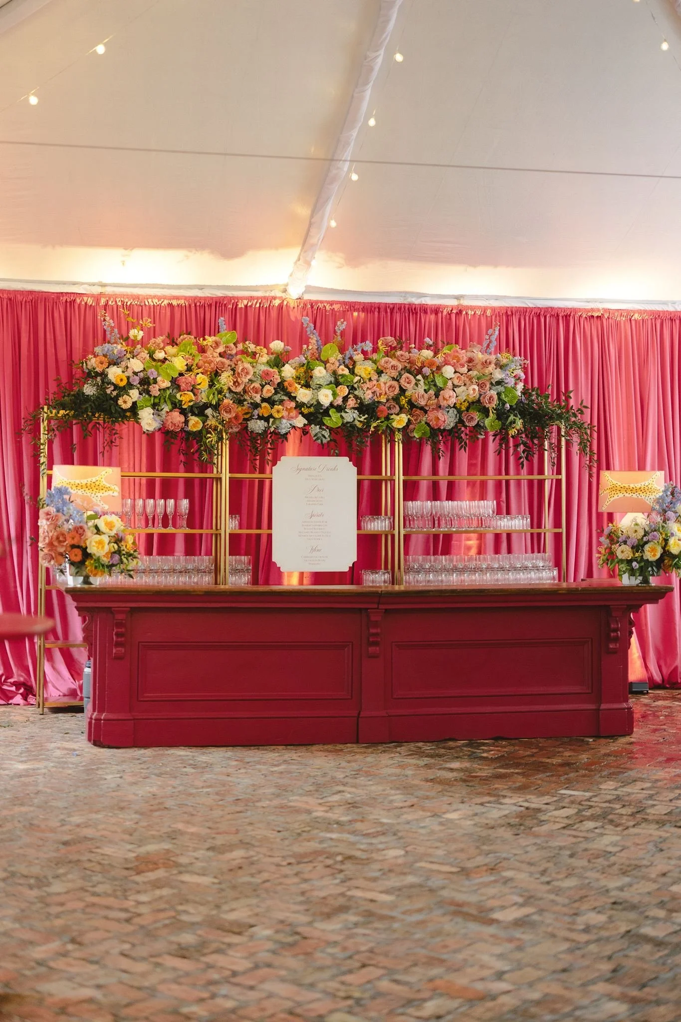 A decorative bar setup with a pink curtain backdrop, floral arrangements, and an elegant menu card in the center.