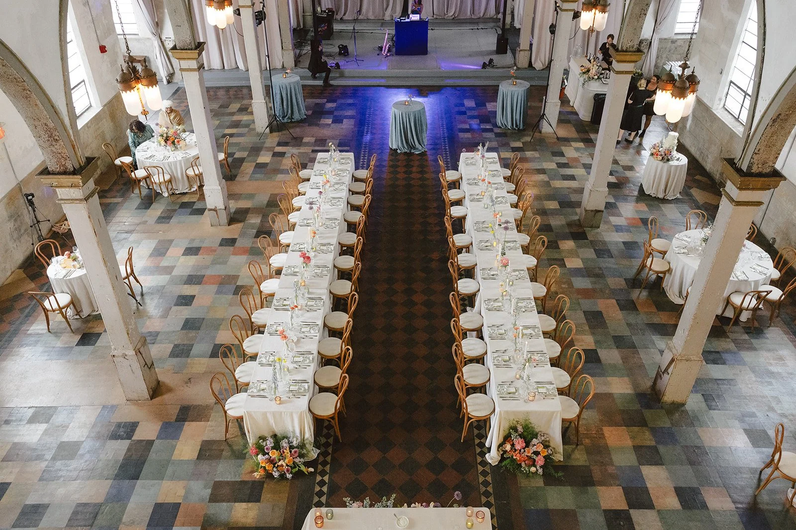A large event hall with high ceilings, tall windows, and vintage-style lighting fixtures. The hall is decorated for a formal event with multiple round and rectangular tables, each covered with white tablecloths and floral centerpieces. A long rectang