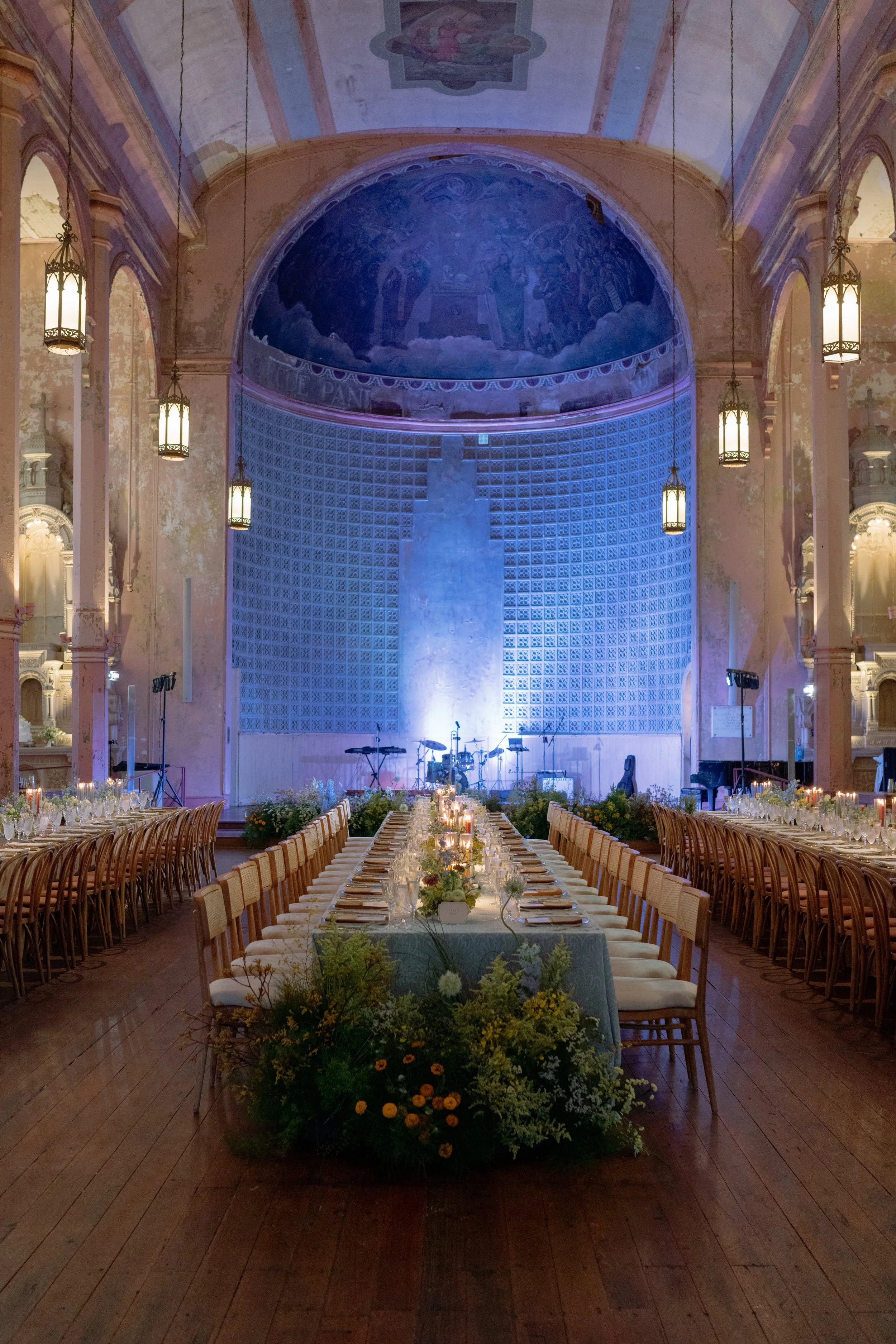 Elegant banquet hall with long dining table decorated with flowers, candles, and place settings, surrounded by wooden chairs. The hall features ornate architecture, high ceilings, and chandeliers, with a stage area at the front and a painted dome cei