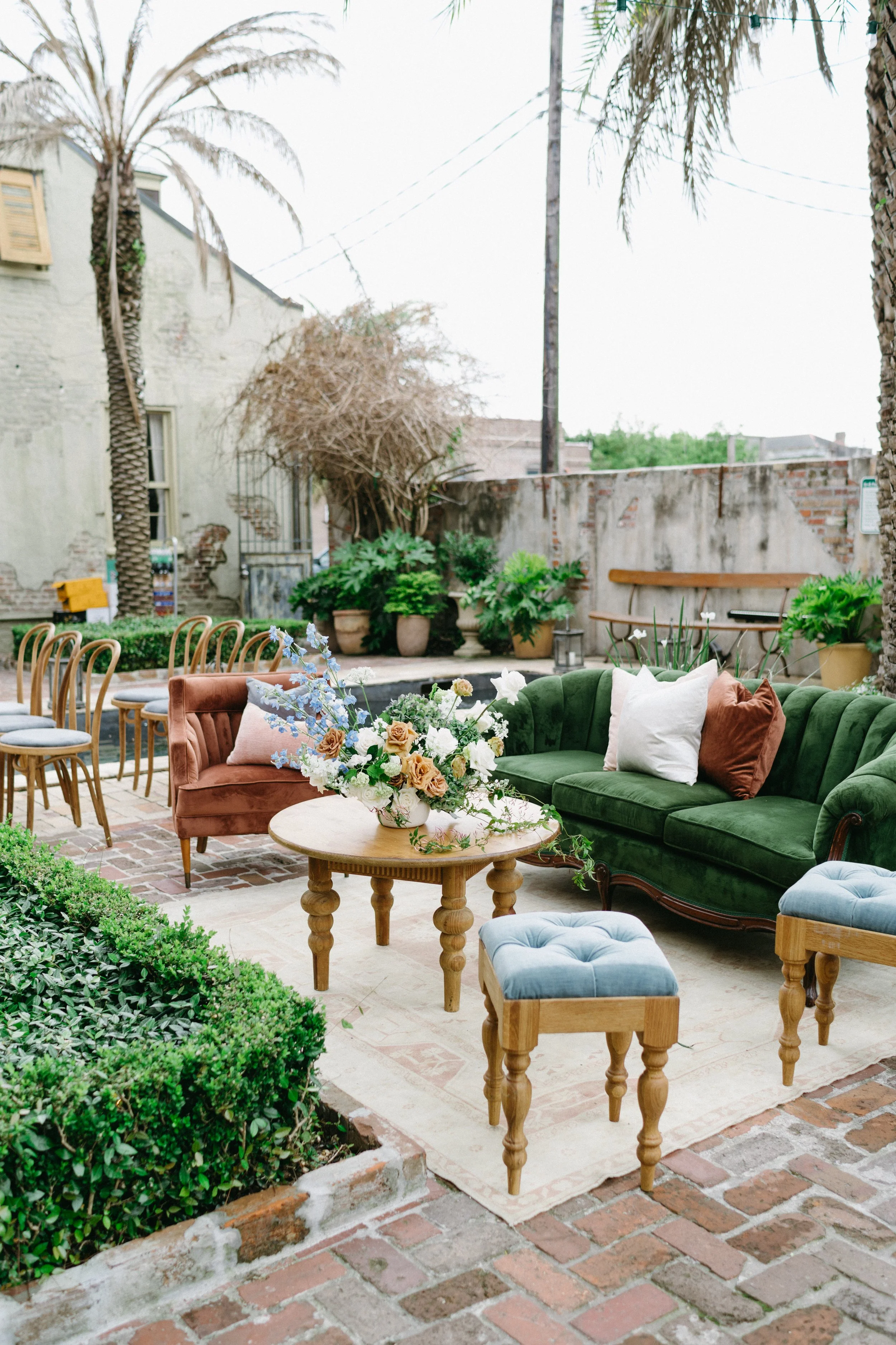 Outdoor patio with vintage and modern furniture, including a green velvet sofa, pink velvet armchair, wooden tables, and chairs, surrounded by potted plants and greenery, with brick flooring and a weathered brick wall in the background.