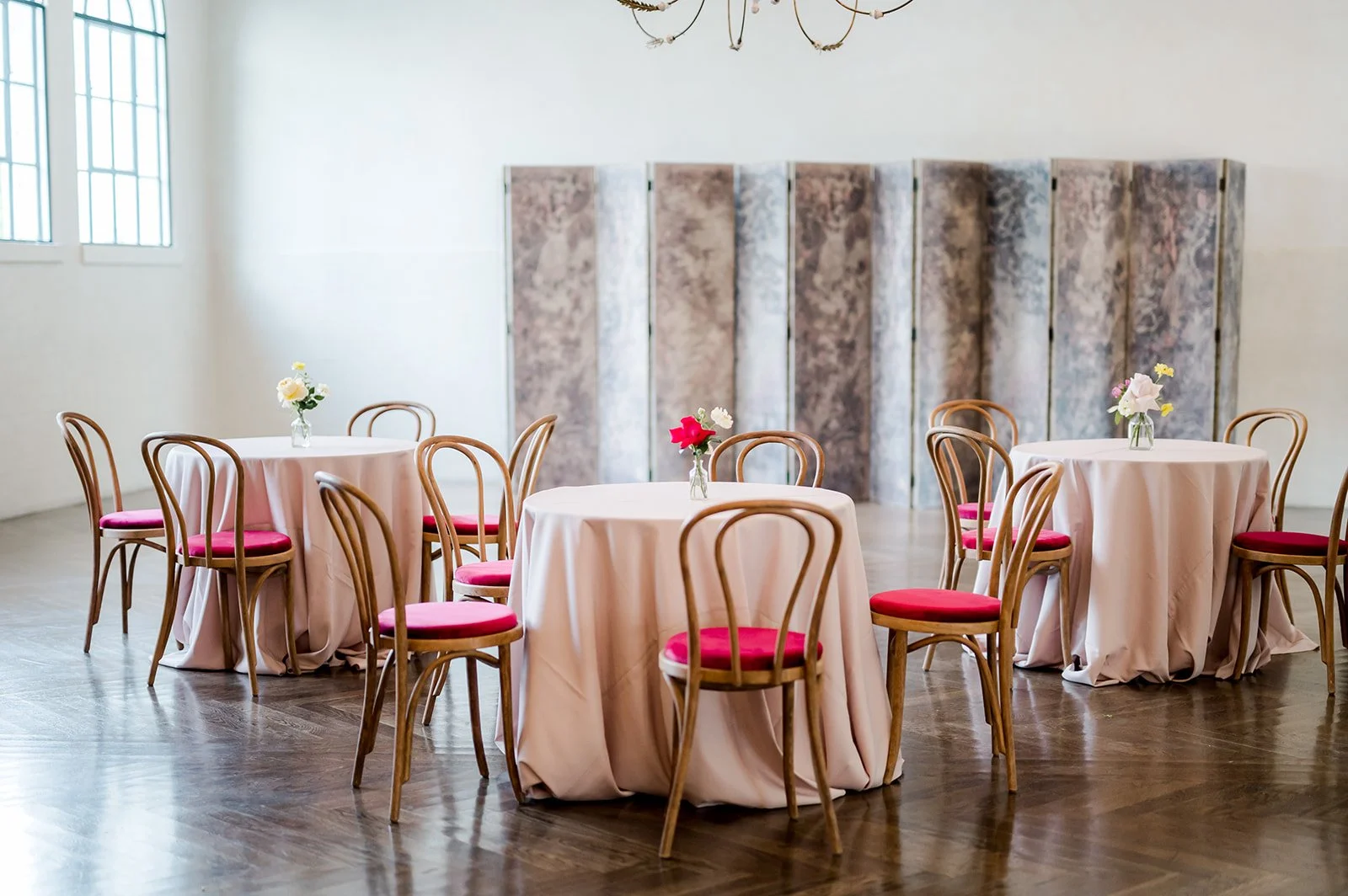An elegant event space with three round tables draped in light pink tablecloths, each decorated with a flower vase containing white, pink, and yellow flowers. The chairs have wooden frames with a rounded back and pink cushioned seats. In the backgrou