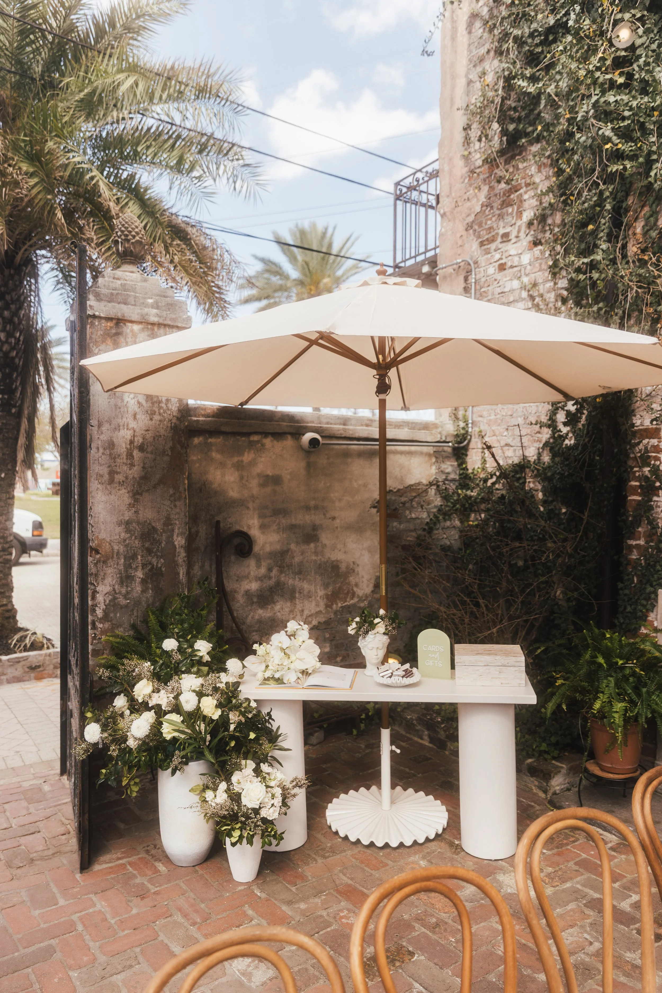 An outdoor table setup with white flowers, decorative items, and a sign on a white table underneath a large white umbrella, with brick flooring and greenery in the background.