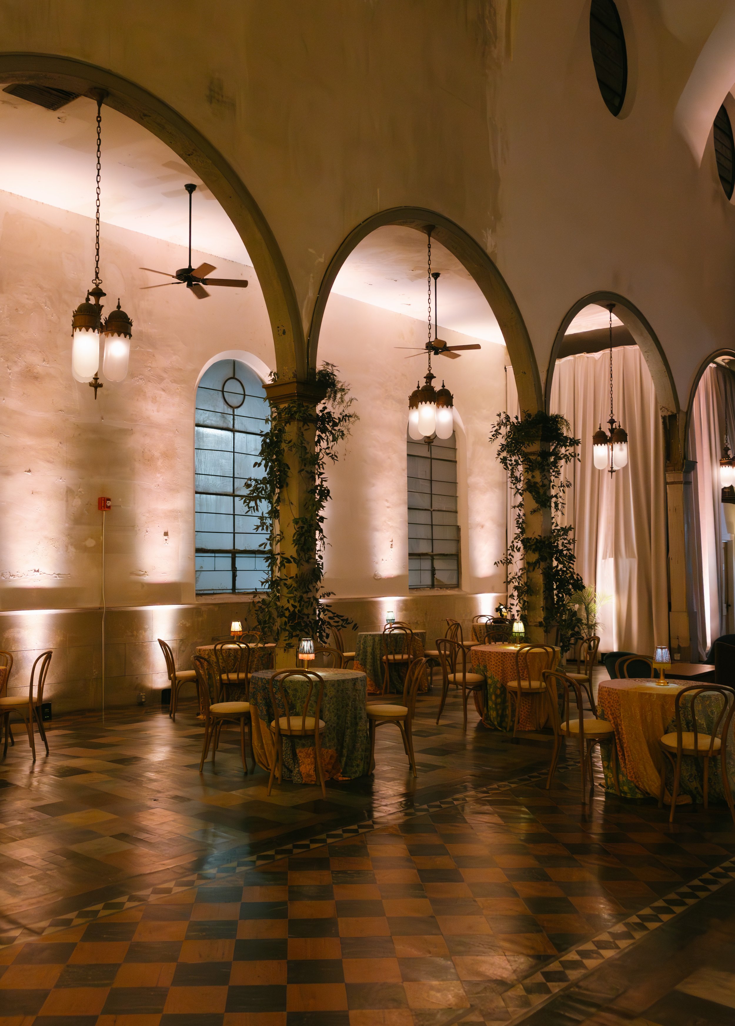 Interior of a restaurant or banquet hall with arched windows, hanging lamps, ceiling fans, and tables with patterned tablecloths and chairs, illuminated by soft lighting.