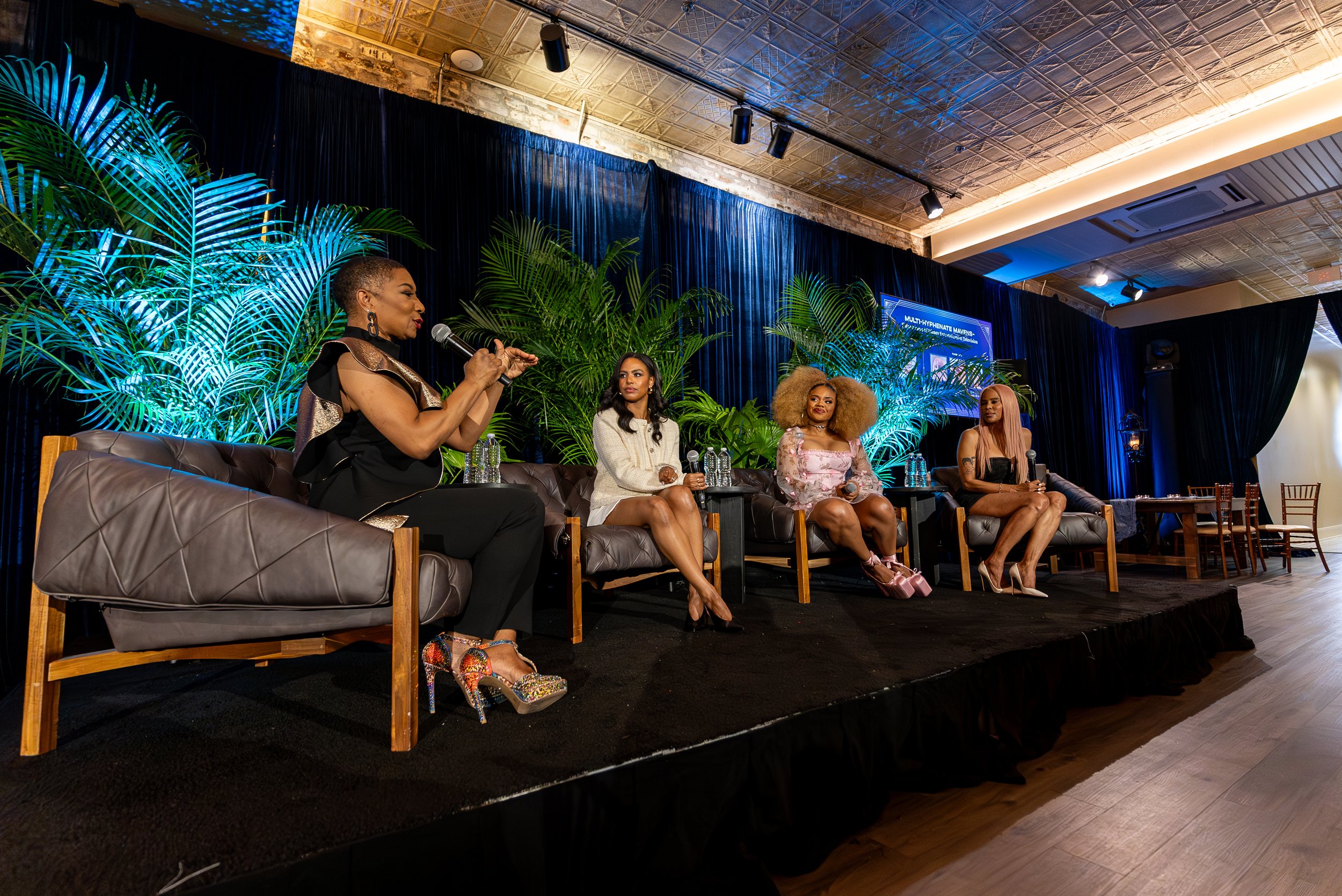 Four women sit on a stage with black curtains and green plants behind them, participating in a panel discussion. One woman is speaking into a microphone, while the others listen. One of the women has colorful high heels. There are water bottles on the table in front of them.