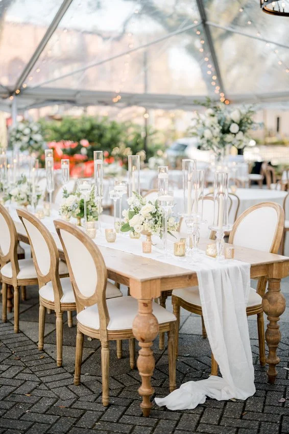 Elegant outdoor wedding reception setup with a wooden table decorated with white flowers and candles, surrounded by cream-colored chairs, under a transparent tent with string lights.
