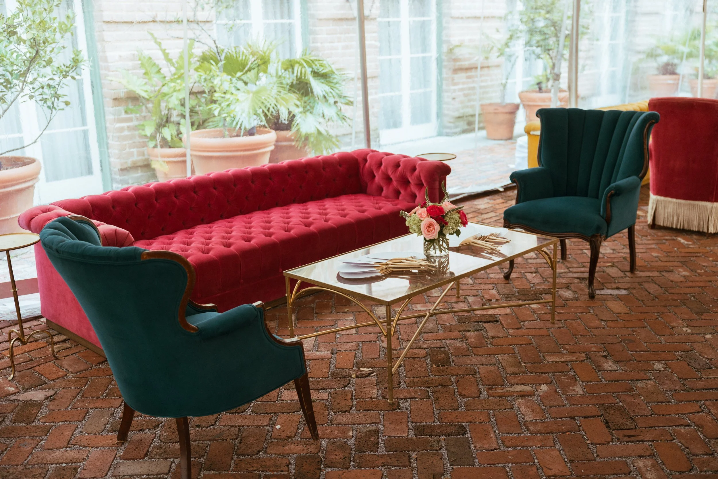 A cozy indoor sitting area with red and green velvet armchairs, a red velvet tufted sofa, a glass coffee table with a flower arrangement, brick flooring, and large windows with potted plants outside.