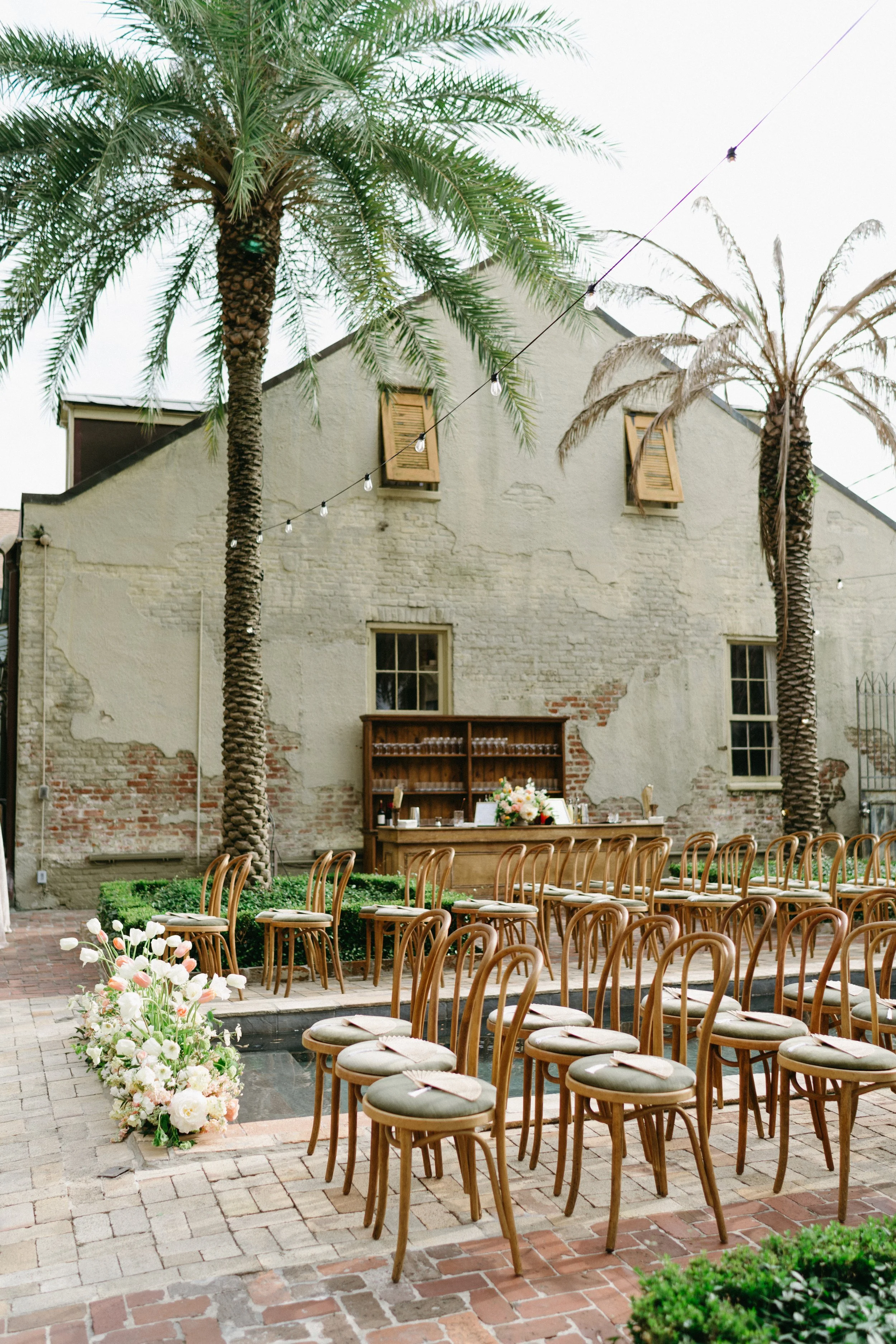 Outdoor venue with wooden chairs arranged in rows, floral arrangements, and a stone and brick building with palm trees and string lights.