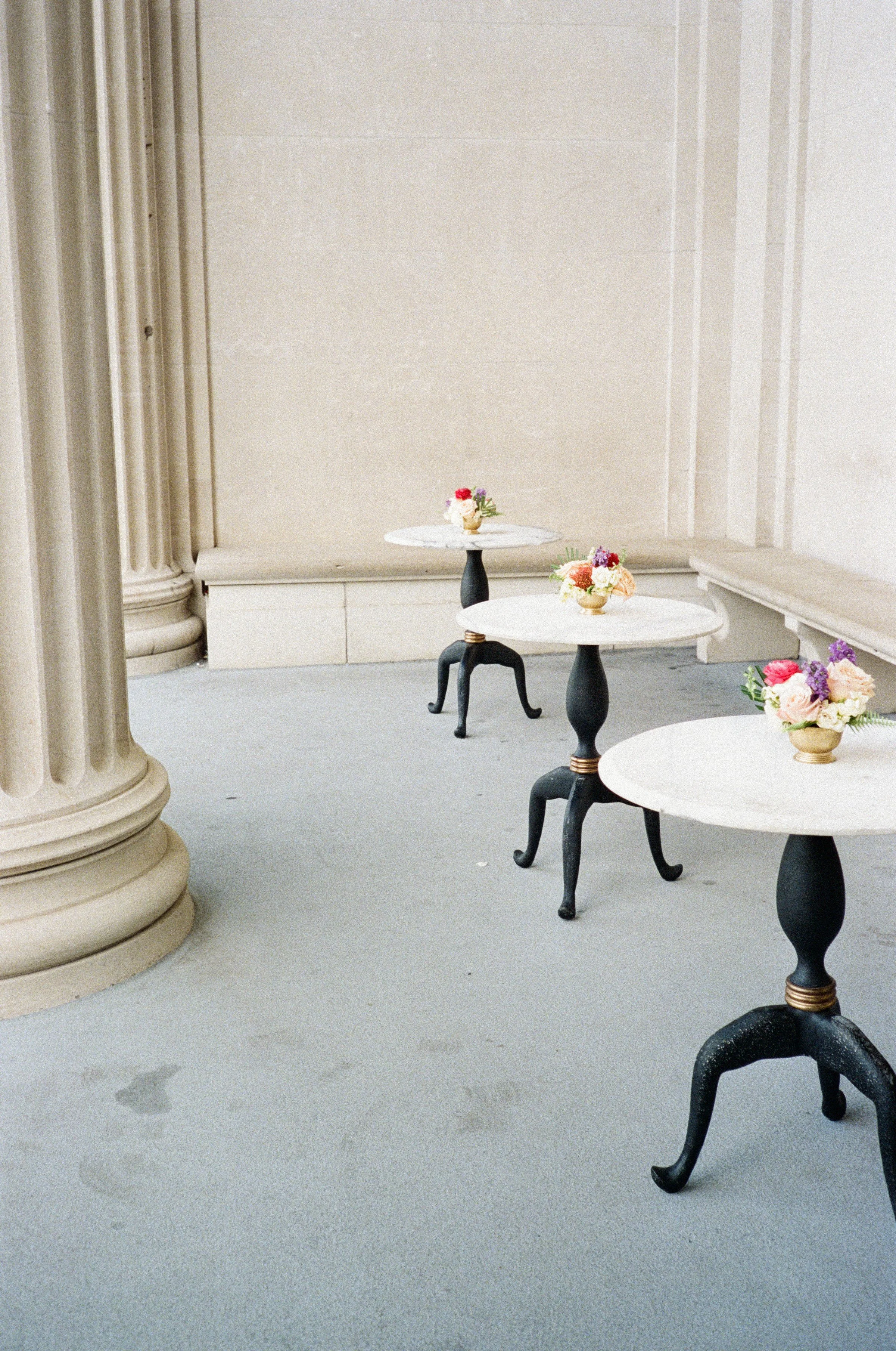 Three small round tables with white tops and black, curved legs, each decorated with colorful flower arrangements, set against a plain beige wall and a long bench along the wall.
