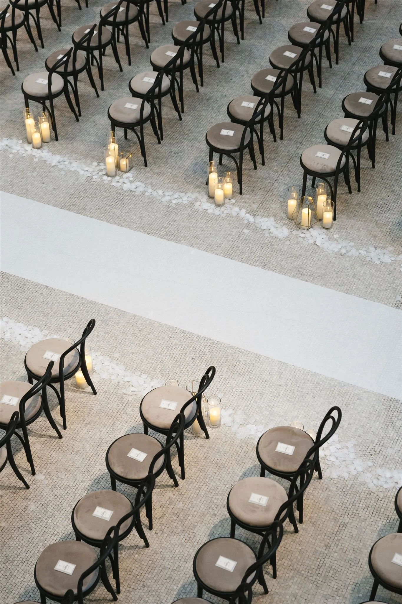 Rows of empty chairs with beige cushions, each with a white paper on top, arranged in a pattern with small candles and white flower petals on the floor underneath in a large hall or event space.