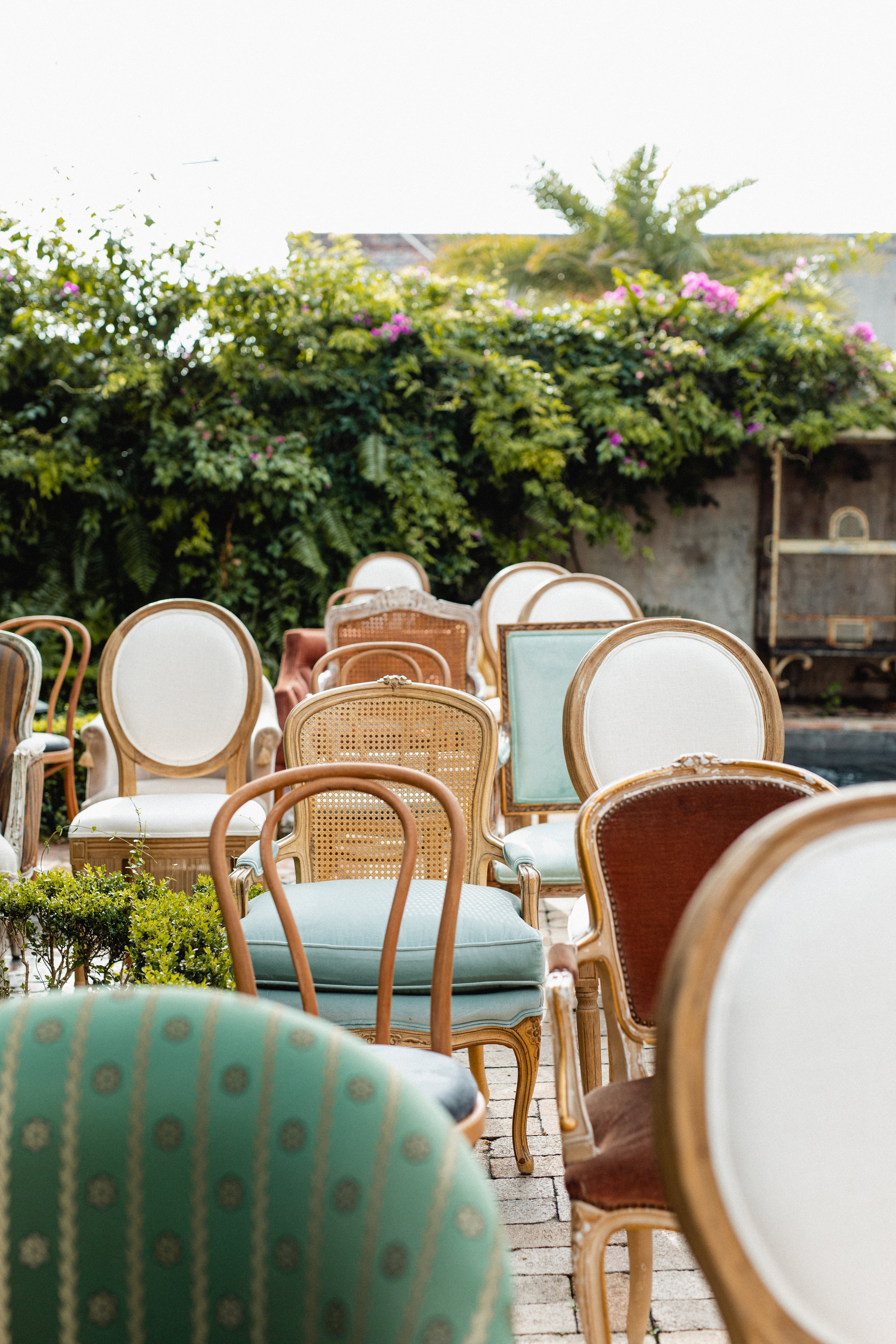 Empty vintage chairs with cushions arranged outdoors on a patio, with greenery and pink flowers in the background