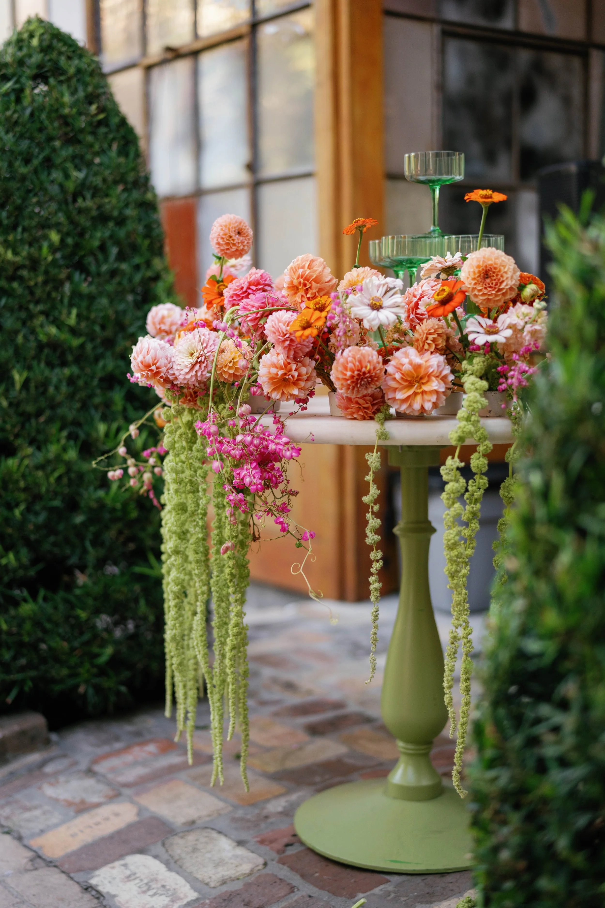 A table decorated with a variety of pink, orange, and white flowers, some hanging down from the table, with a green glass tiered cake stand on top, placed outdoors on a brick patio near green bushes and a wooden structure.