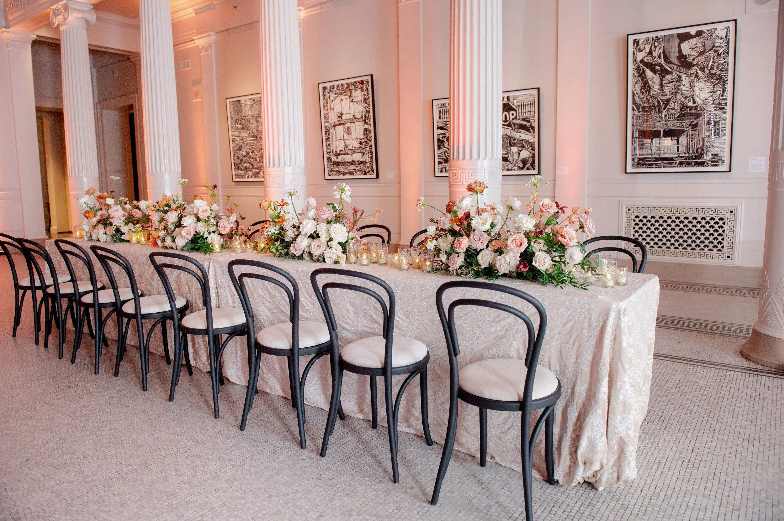Elegant event space with long table decorated with pink and white floral arrangements and lit candles, surrounded by black and white chairs, with framed artwork on the walls and white columns.