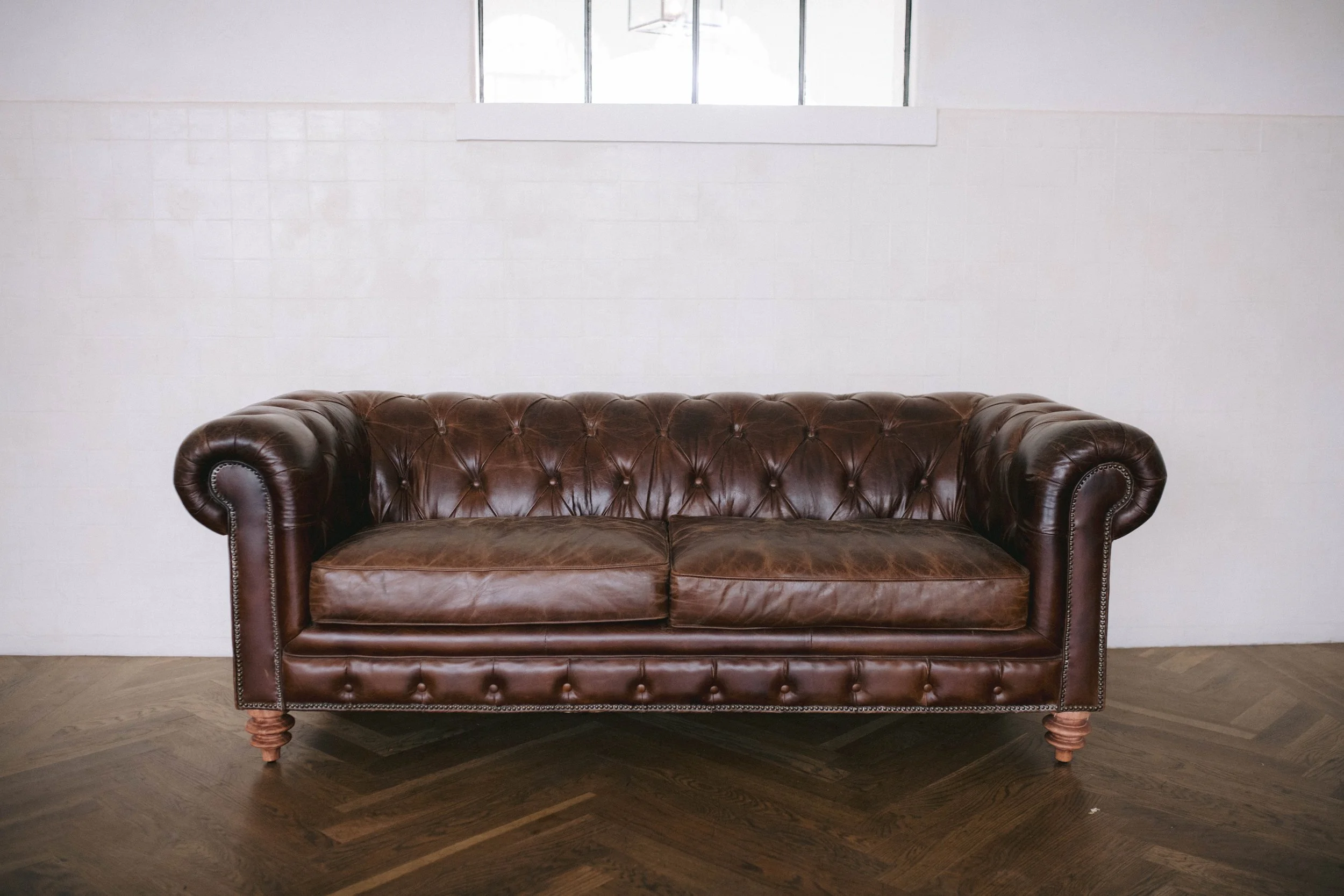 A vintage brown leather Chesterfield sofa with tufted design, rolled arms, and wooden legs situated on a dark hardwood floor against a plain white wall with a small rectangular window above.