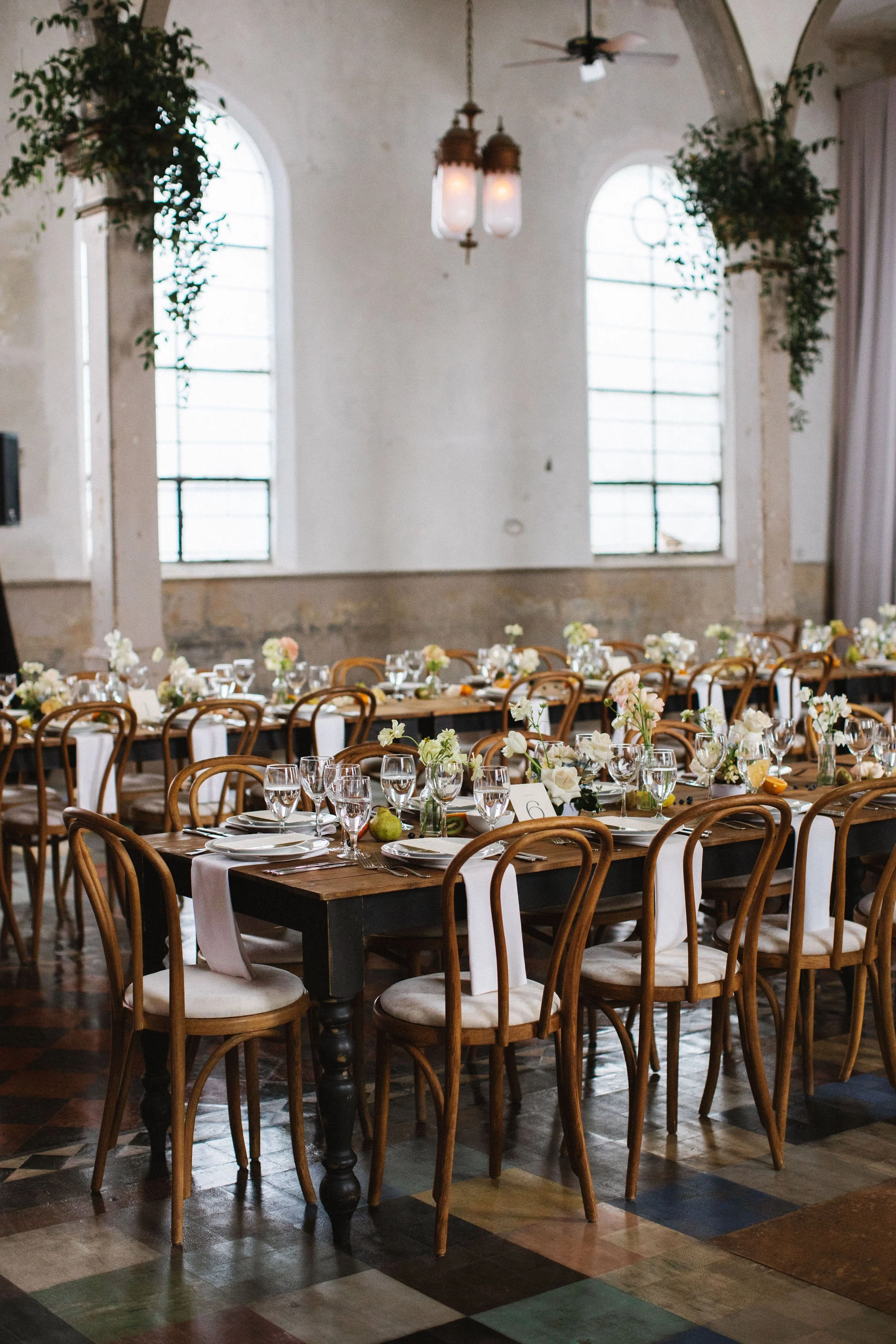 A decorated banquet table set for a formal event with floral centerpieces, glassware, and plates inside a rustic, vintage-style room with arched windows and hanging lamps.