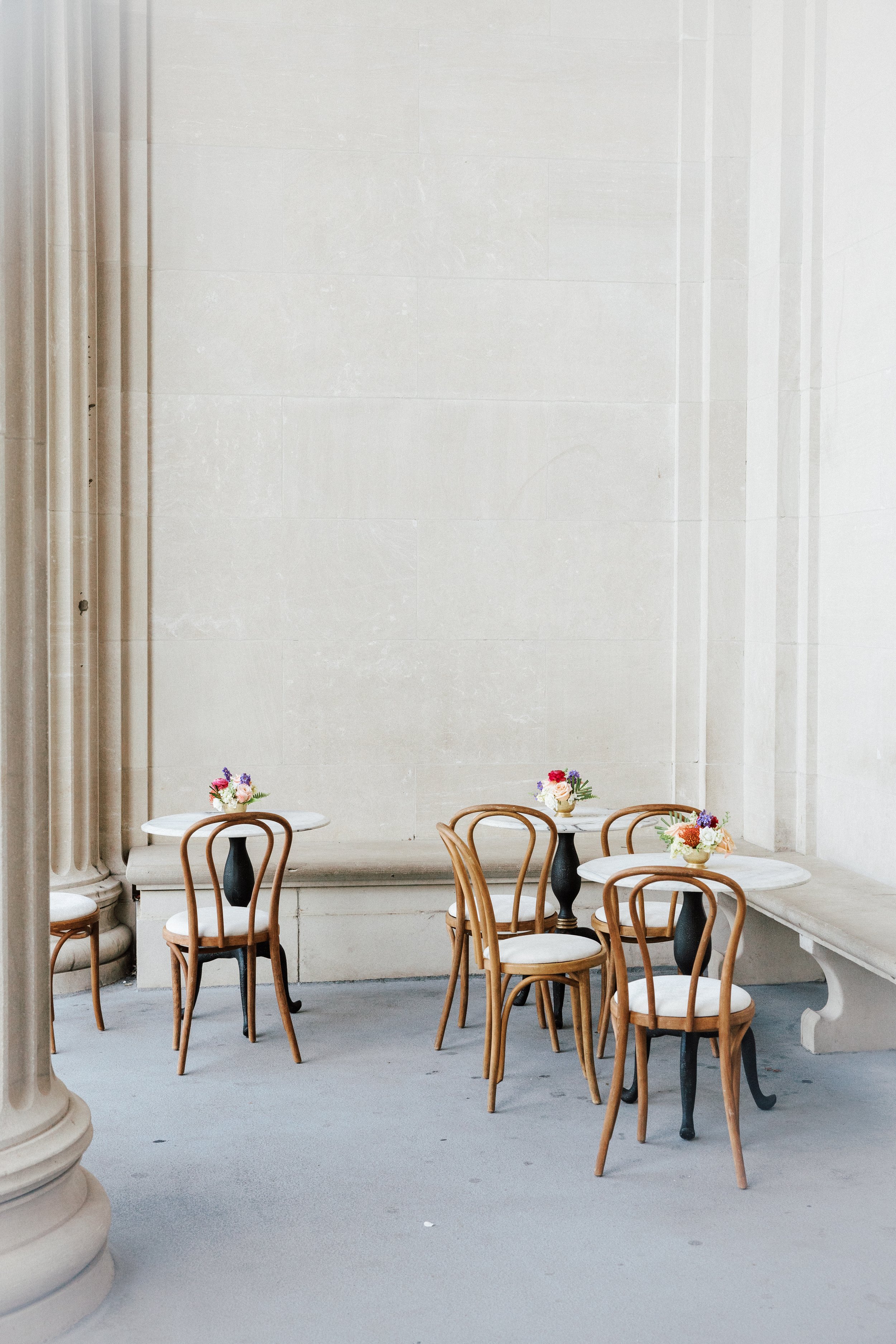 Indoor seating area with three round tables decorated with small flower arrangements, surrounded by wooden chairs with white cushions, against a white wall with a classical architectural column on the left.