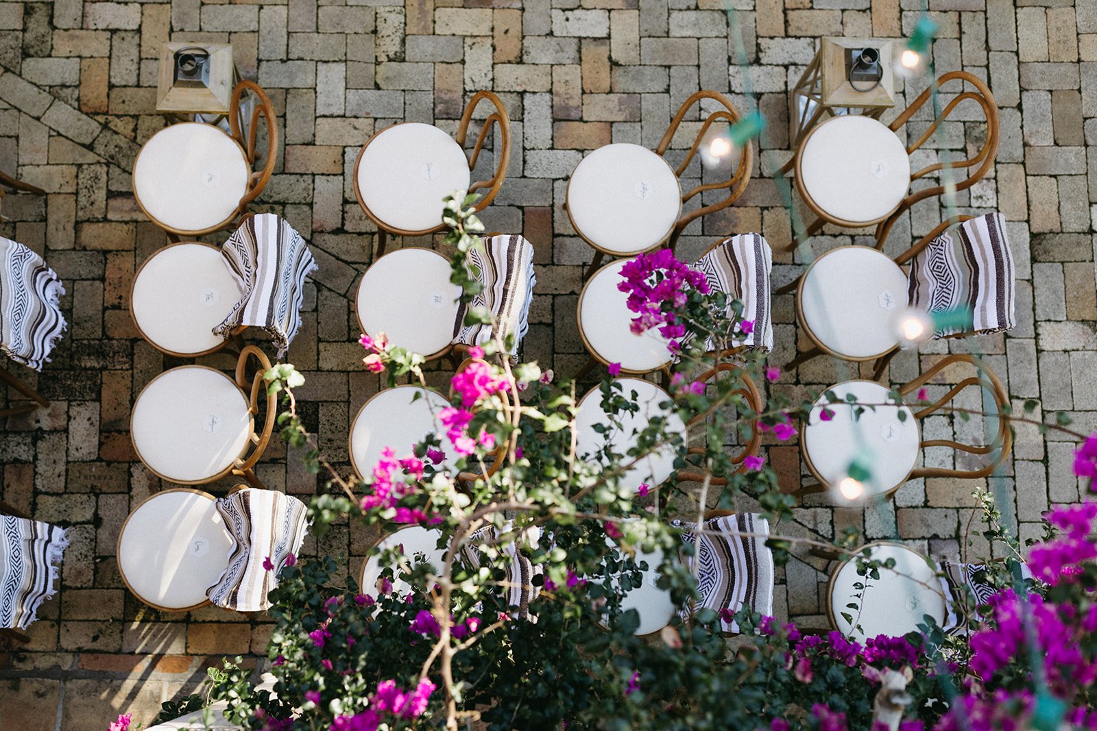 Overhead view of white round tables with striped cloths and wooden chairs on a brick patio, decorated with pink bougainvillea flowers and string lights.