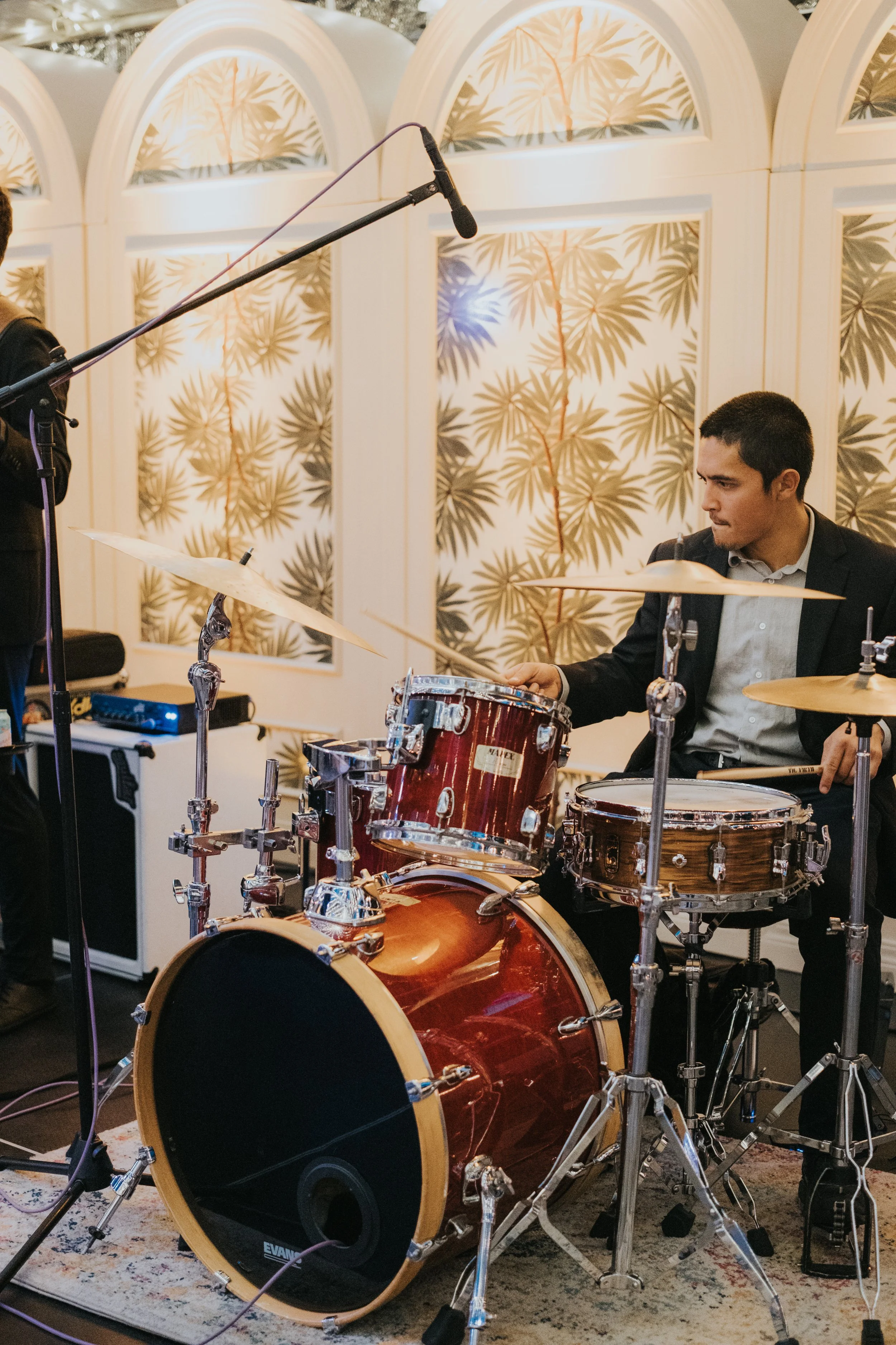 A man in a suit playing a drum set at a formal event with decorative wallpaper in the background.